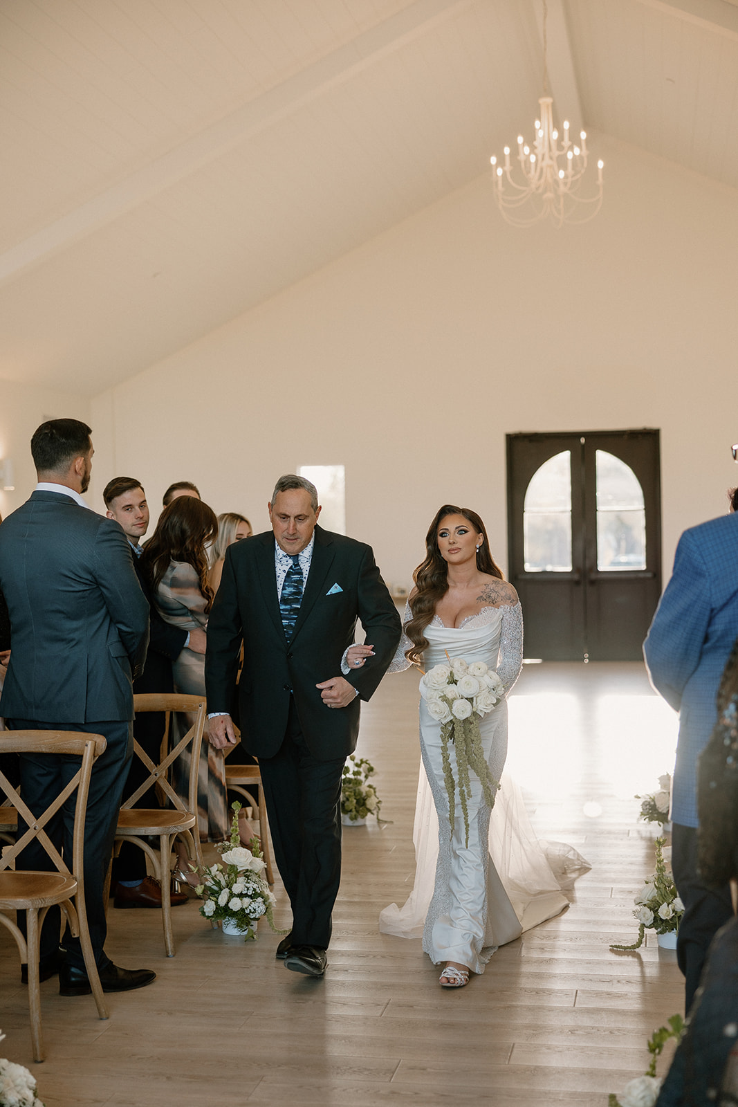 Bride walks arm-in-arm with her father down the aisle, surrounded by loved ones and florals inside the elegant white chapel at a desert view wedding venue.