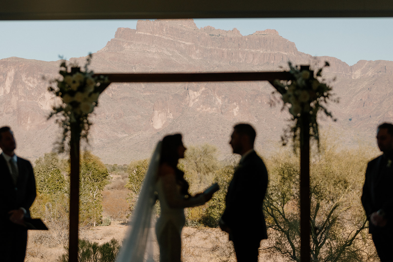 Silhouettes of the bride and groom exchanging vows under a wooden arch, framed perfectly by towering mountains behind them at a desert view wedding venue.