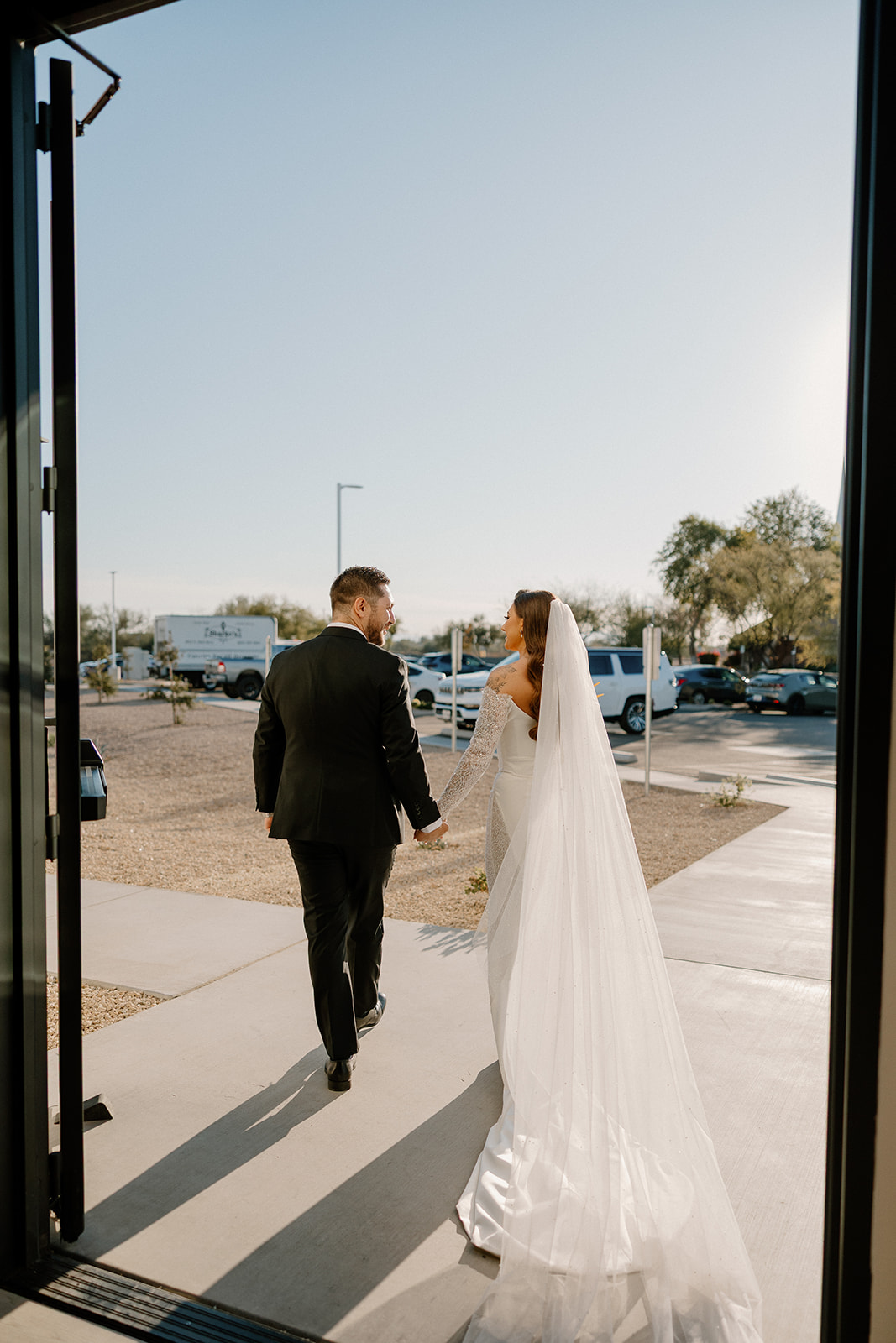 Bride and groom walk through the open doors of their venue, holding hands as sunlight pours across the pavement behind them.