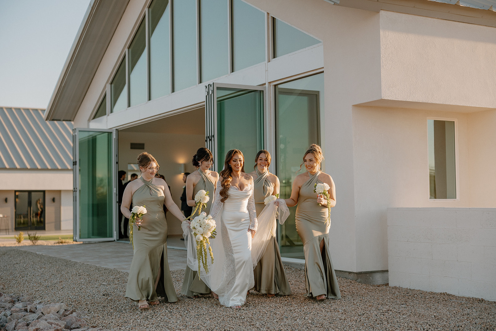 Bride and her bridesmaids walk together outside the modern venue, holding white florals with the late afternoon light catching their satin gowns at a desert view wedding venue.