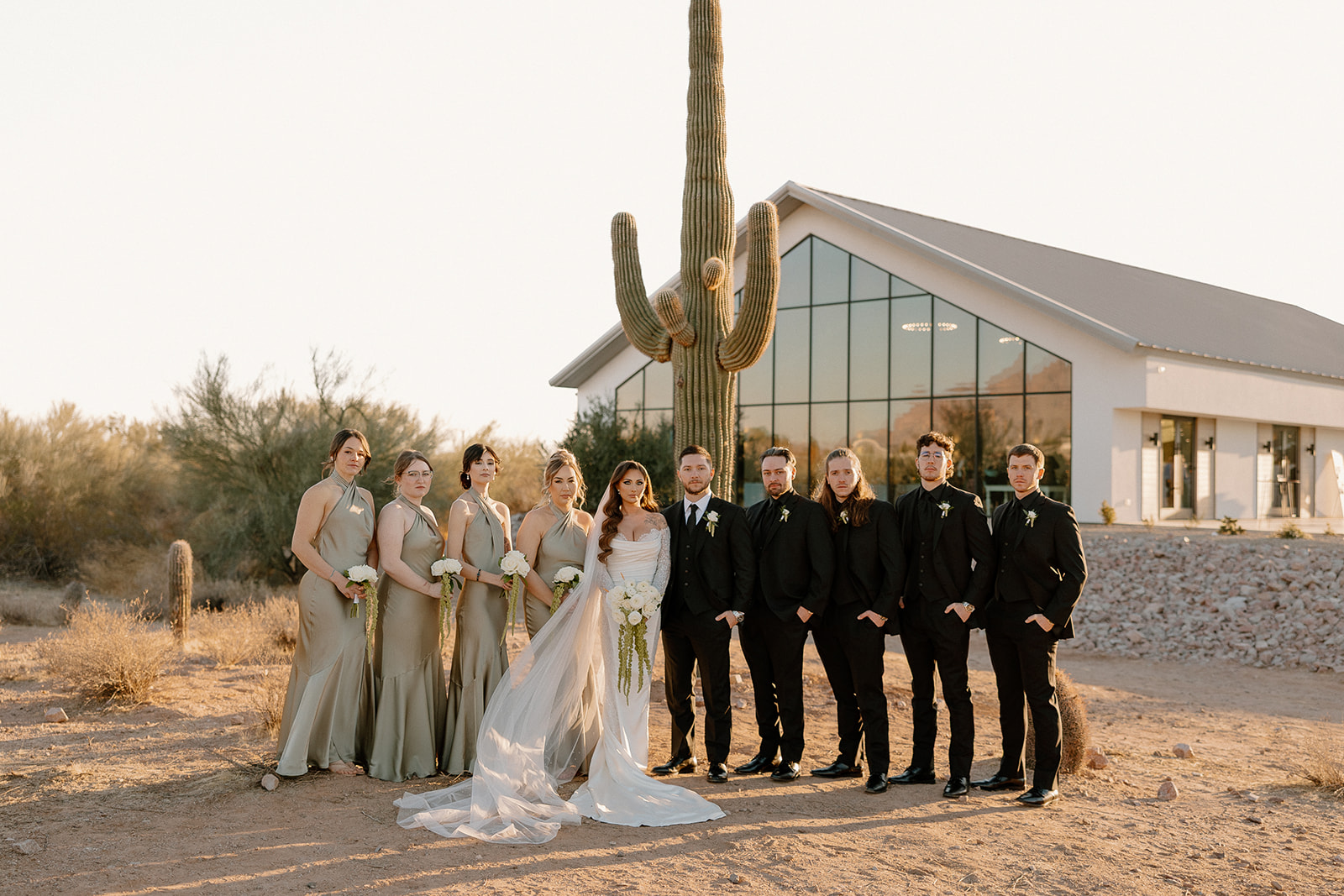Bride and groom stand with their wedding party in neutral tones, framed by a towering cactus and the glass-fronted chapel of their desert view wedding venue.