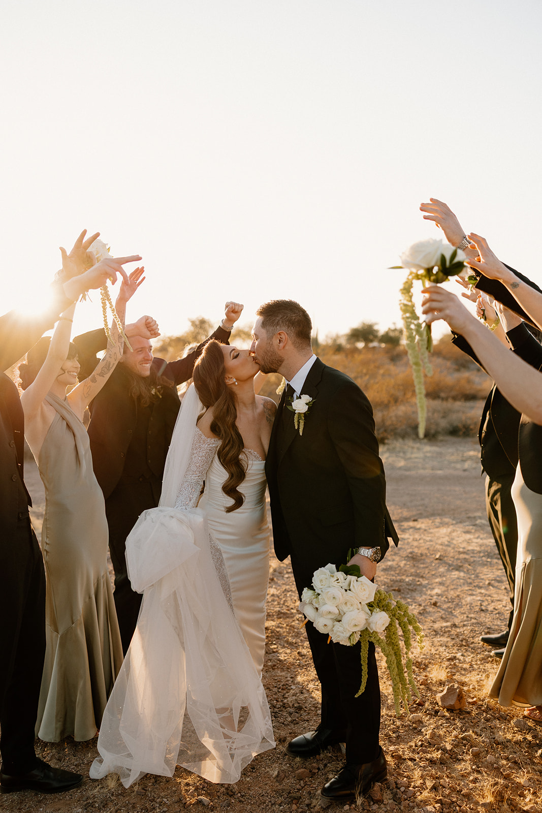 Bride and groom kiss while walking through an arch of raised hands from their wedding party, golden light shining behind them.