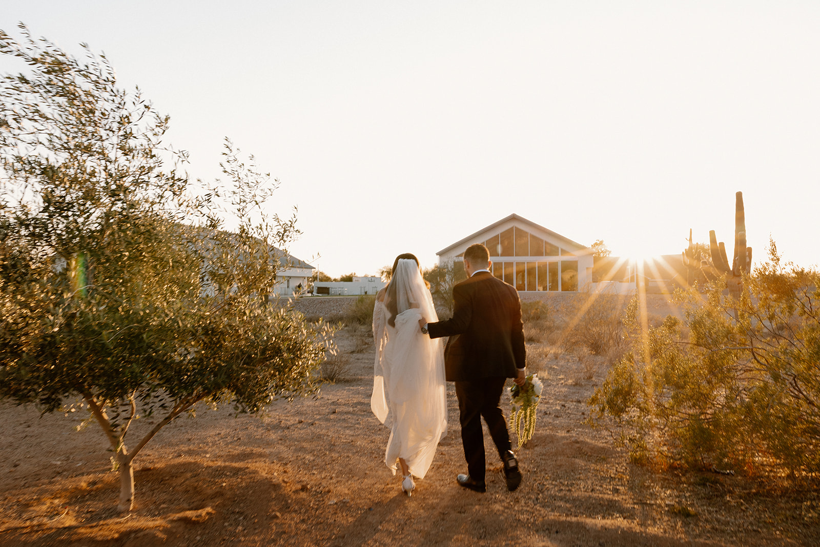 Bride and groom walk through the desert as the sun sets behind the chapel at their desert view wedding venue, casting long shadows and warm glow.