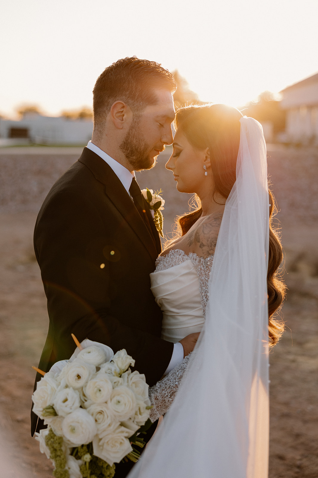 Groom holds bride close as the sun flares behind her veil, white rose bouquet in hand and desert tones in the background.
