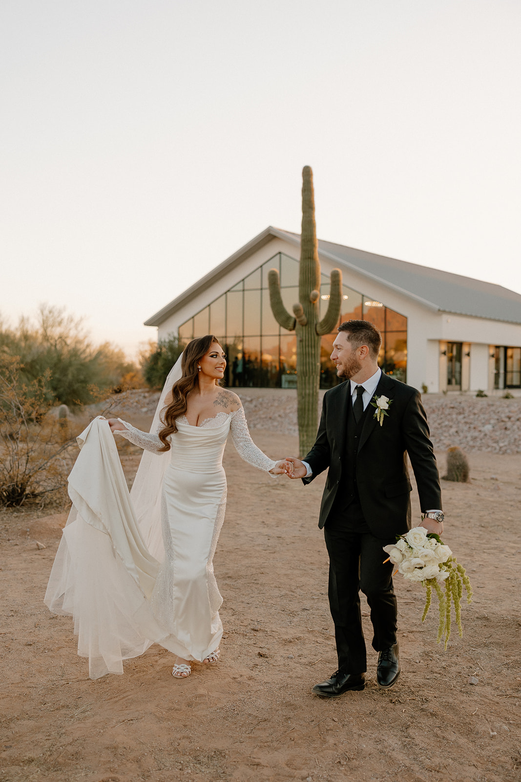 Bride and groom share a soft laugh while walking hand-in-hand through the desert terrain, their venue and a towering cactus behind them at a desert view wedding venue.