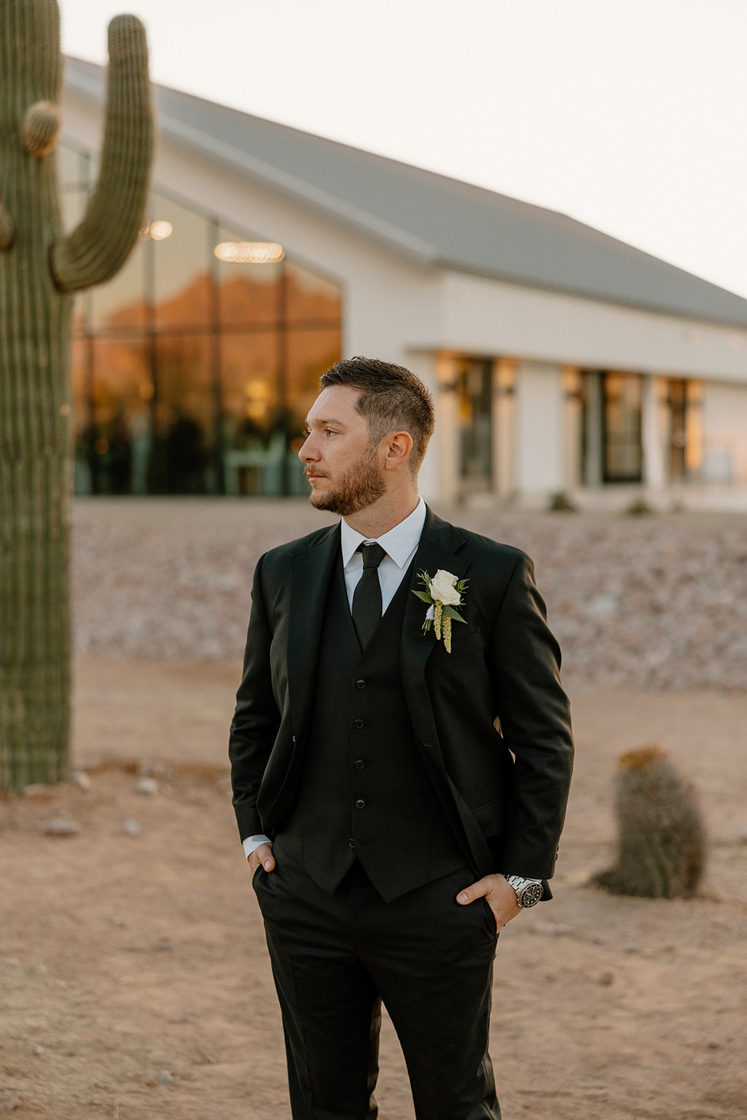 Groom stands in a black suit outside a modern white chapel, desert terrain and a tall cactus setting the Arizona scene.