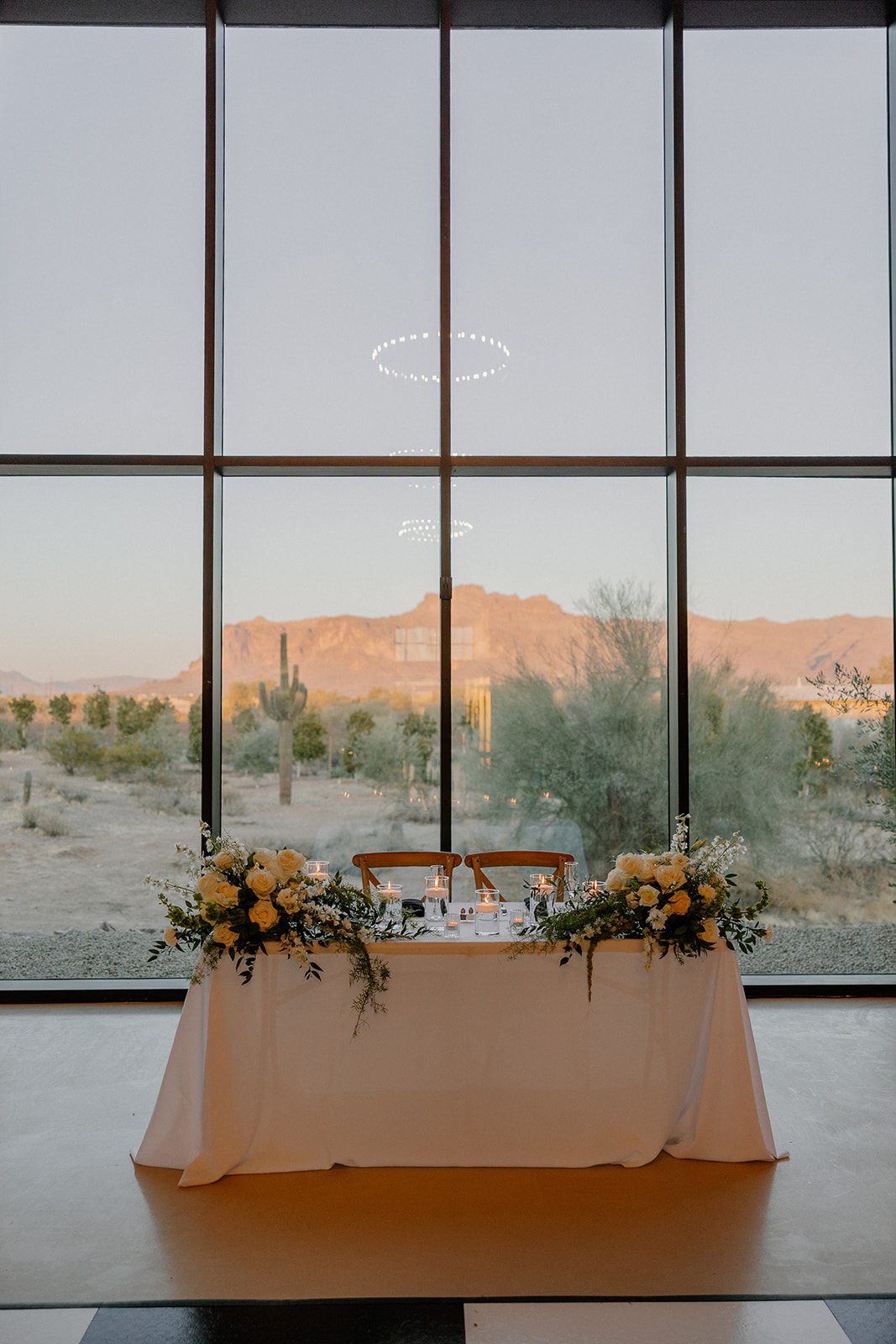 Sweetheart table framed by floor-to-ceiling glass, perfectly centered with Superstition Mountains visible behind it at the desert view wedding venue.