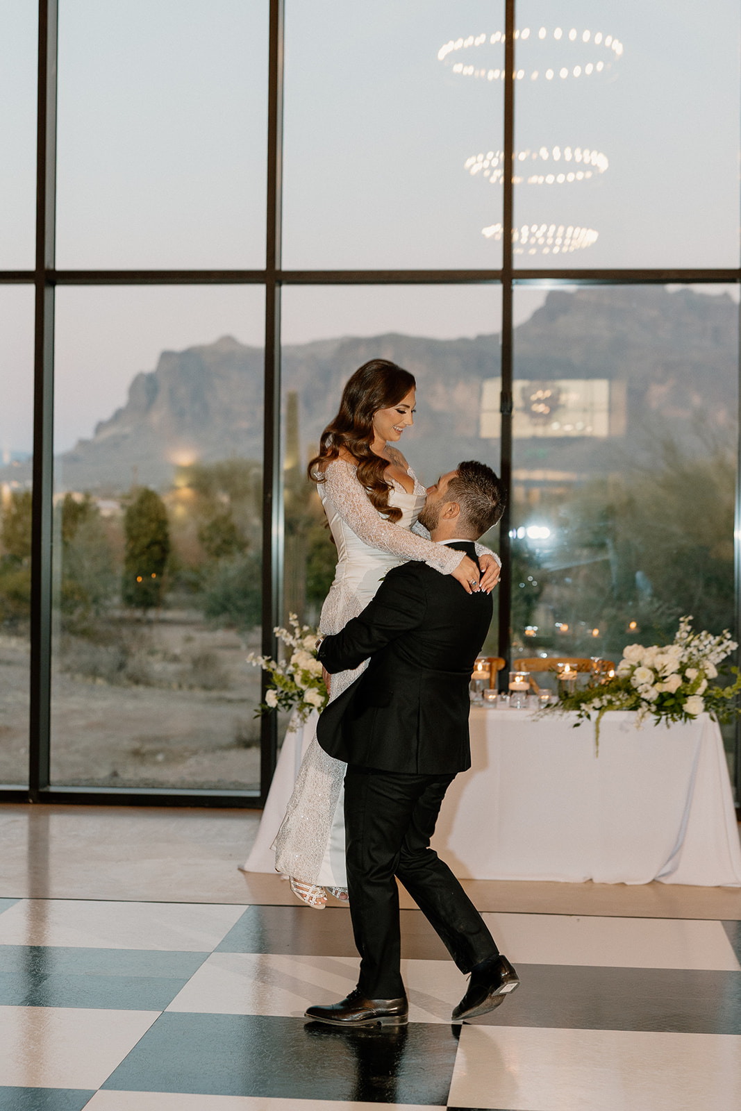 Groom lifts the bride during their first dance on a black-and-white checkered floor, with floor-to-ceiling windows revealing mountain views beyond.