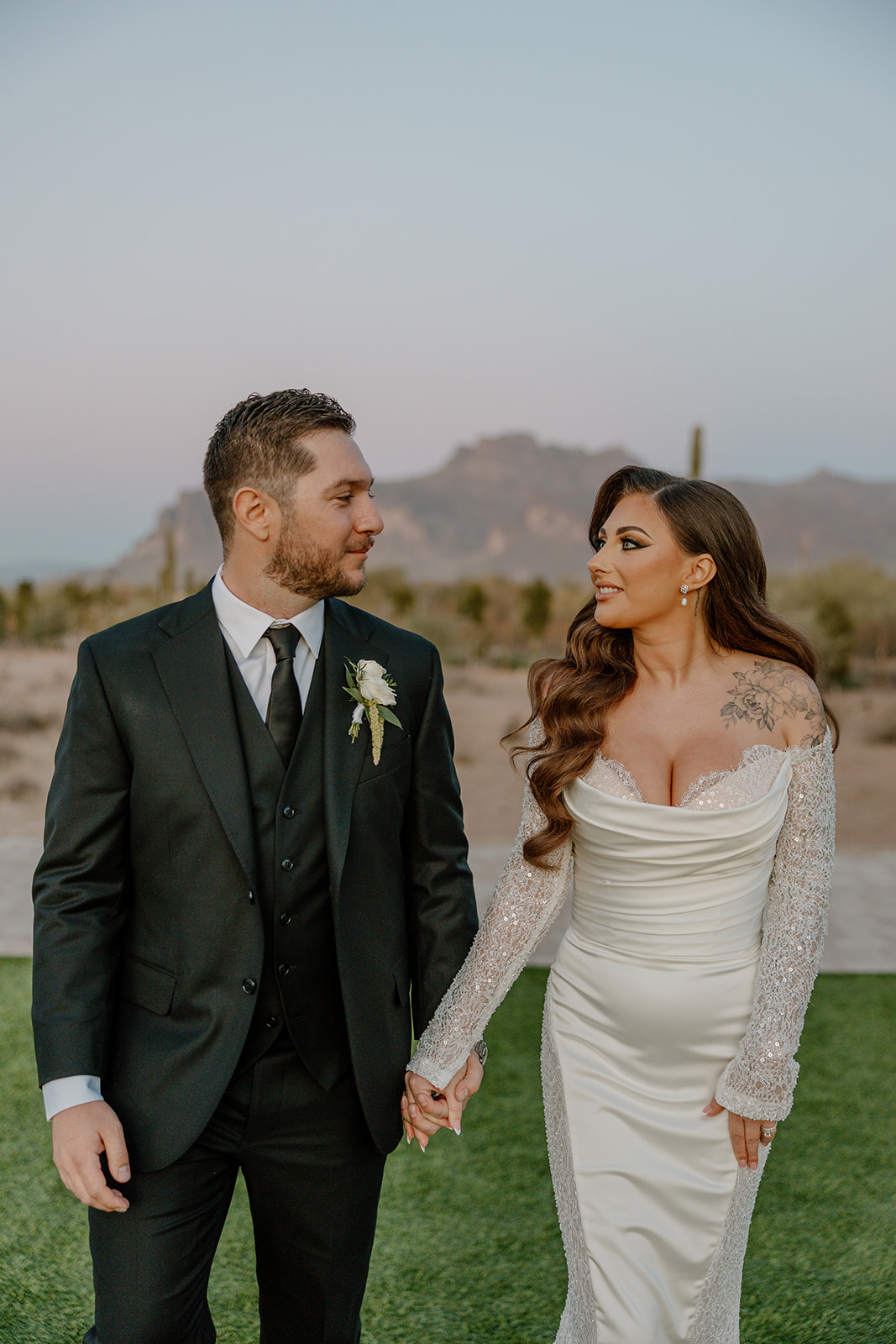 Bride and groom walking hand in hand at golden hour, with rugged mountains and cacti behind them at a desert view wedding venue.