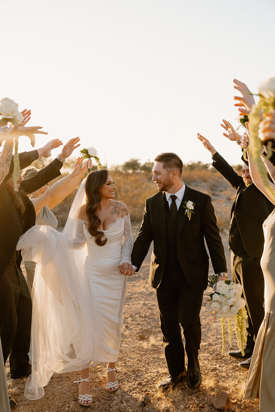 Bride and groom walk through a tunnel of raised hands from their wedding party, glowing in the golden hour light of a desert view wedding venue.