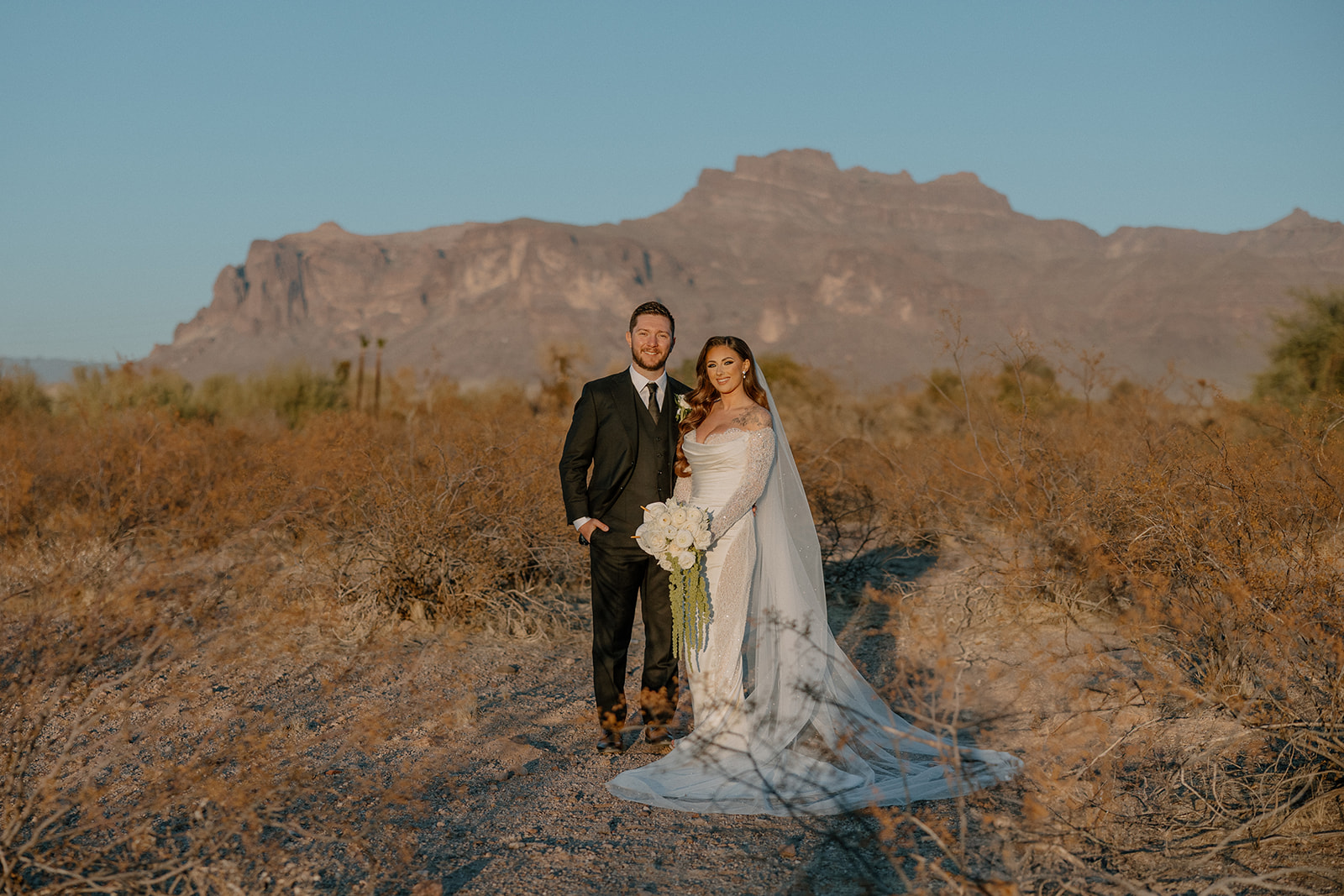 Bride and groom stand hand-in-hand on a desert path, framed by dramatic mountains and golden light at a desert view wedding venue.