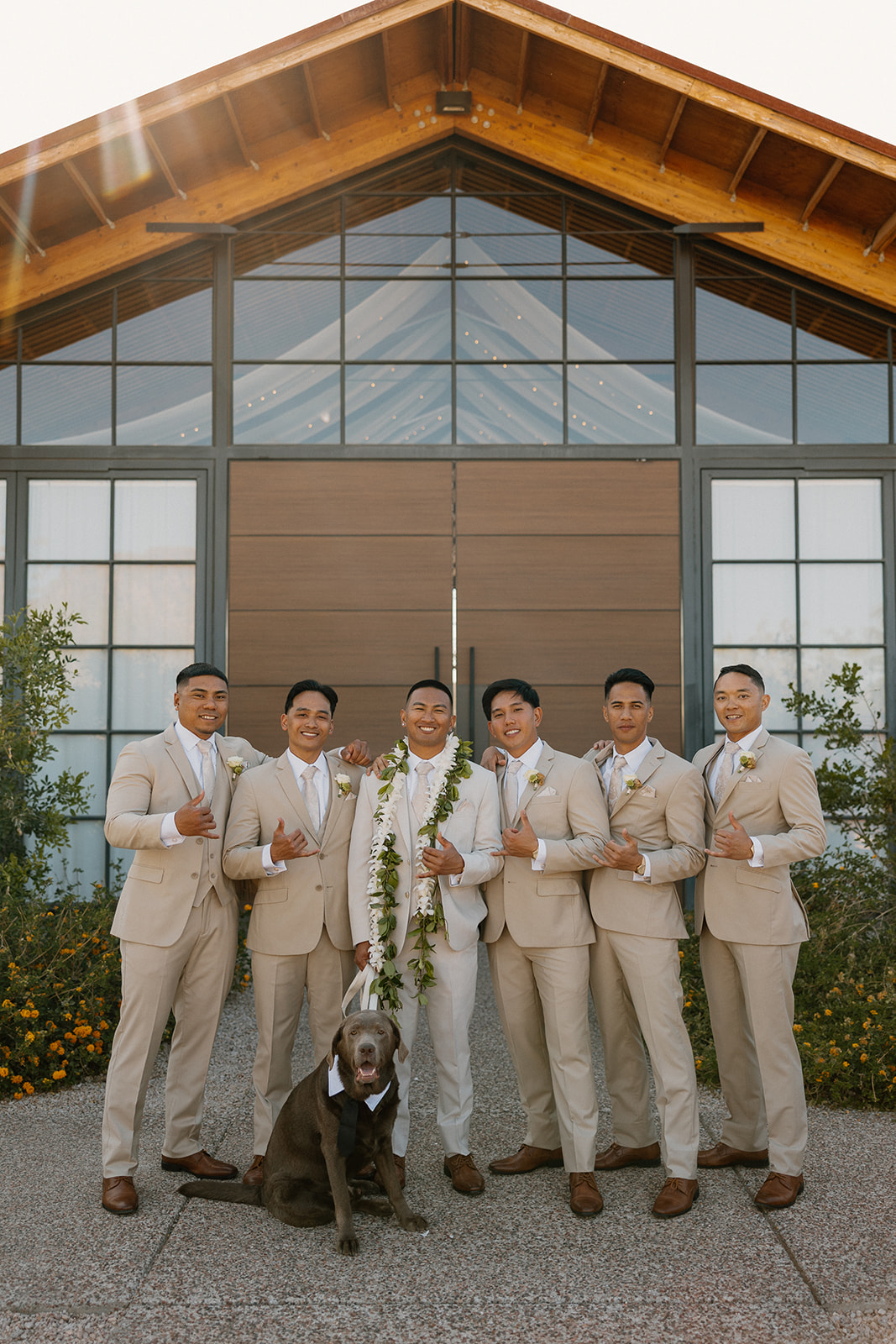 The groom and his groomsmen pose outside the venue with their dog, all in tan suits and looking sharp.