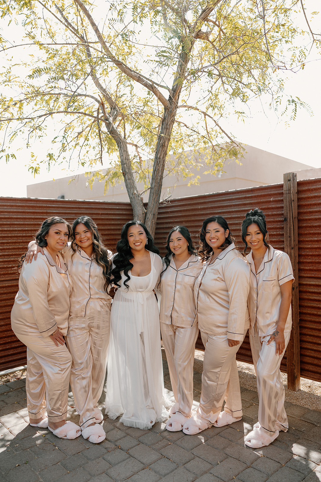 The bride in a white robe with her bridesmaids in champagne pajamas, all smiling under a desert tree before the ceremony.