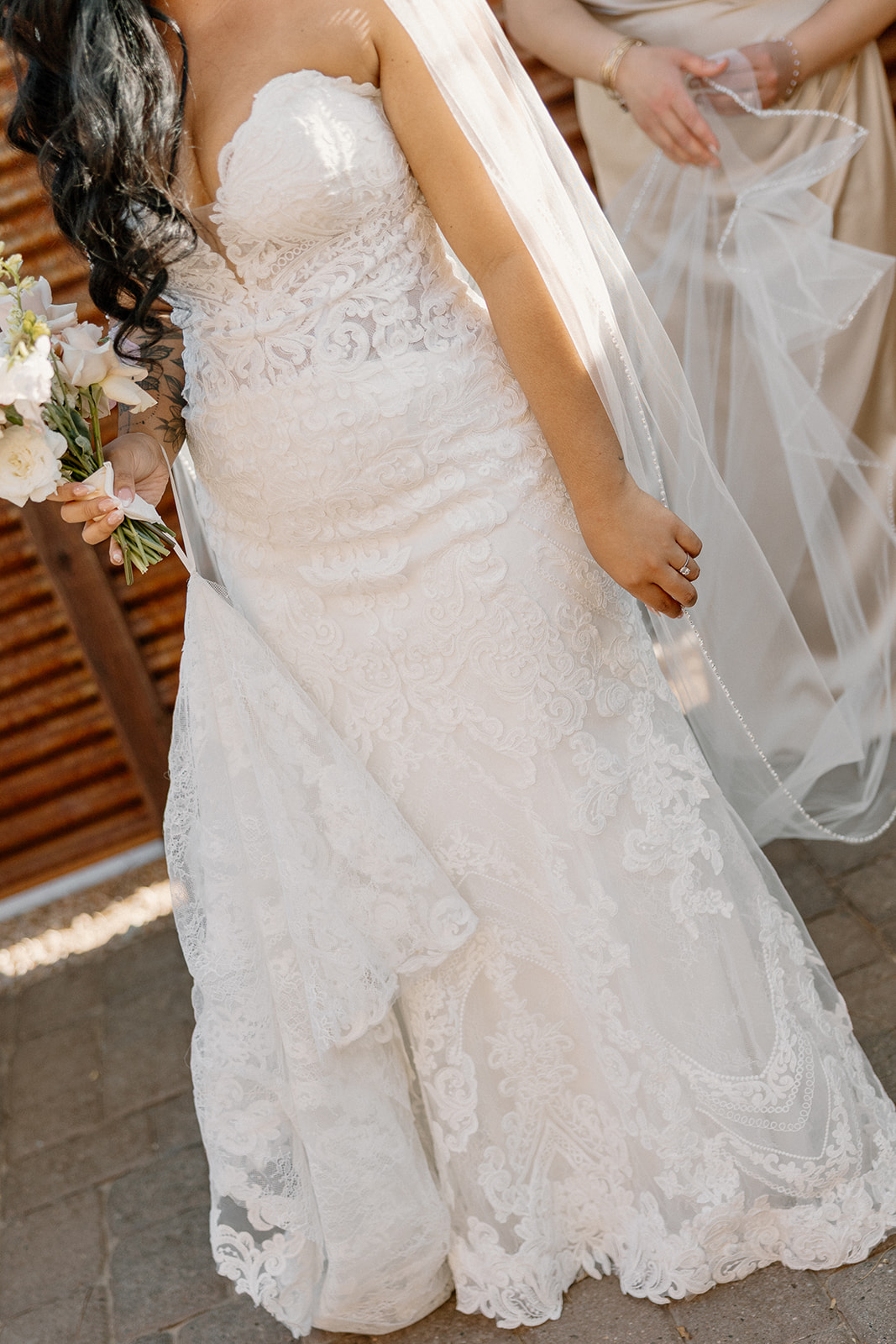 Close-up of intricate lace detail on the bride’s gown as she holds a soft floral bouquet, sunlit and glowing.