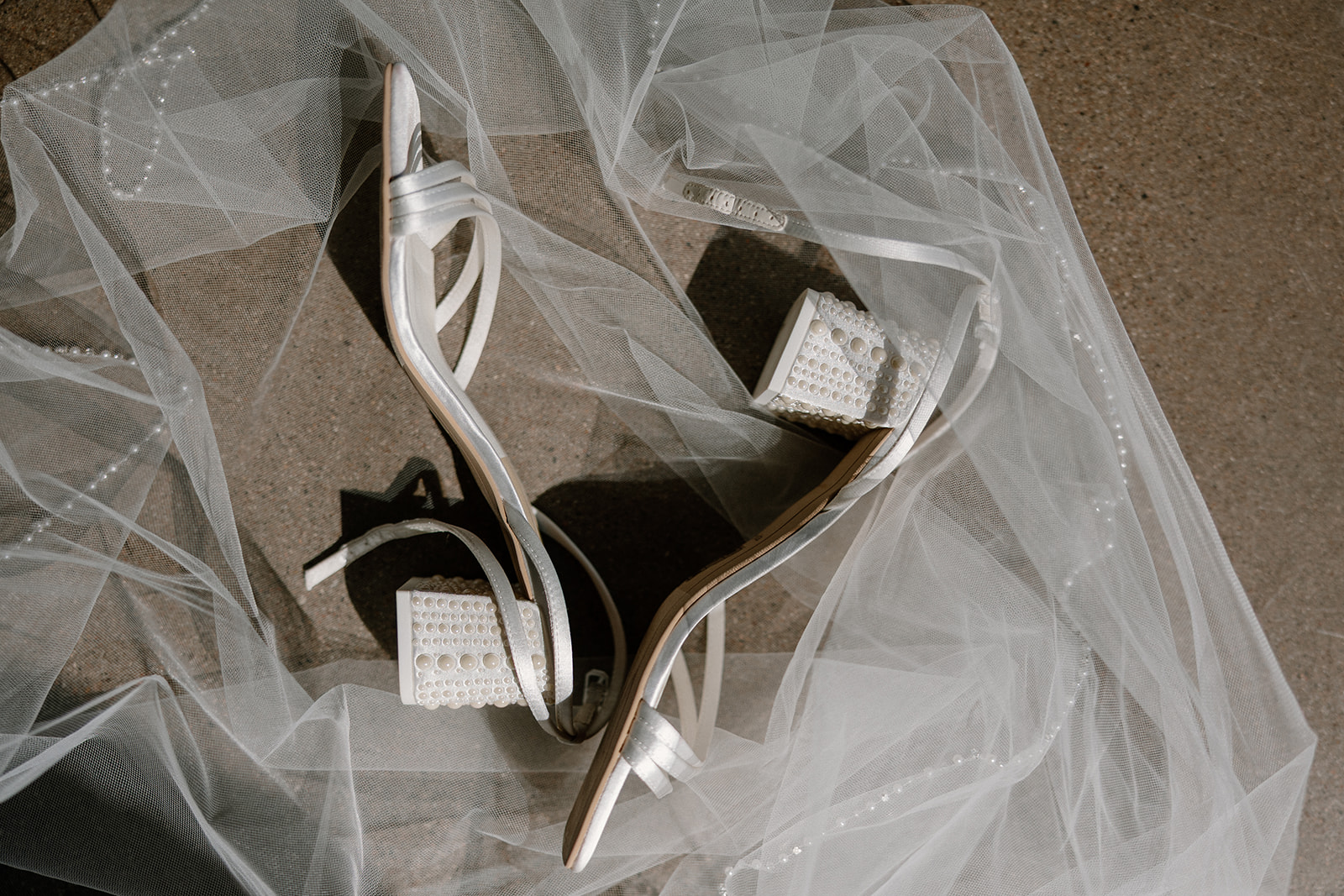 Detail shot of pearl-block heels and bridal veil catching the sunlight on rustic pavers.