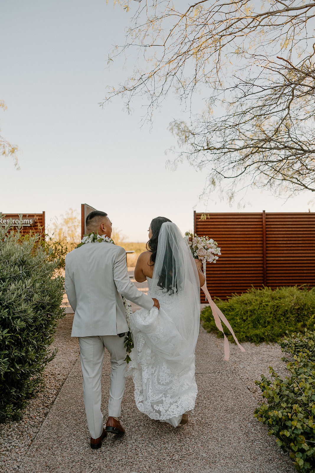 Newlyweds walk side by side down a desert garden path, bouquet in hand and light filtering through the trees.