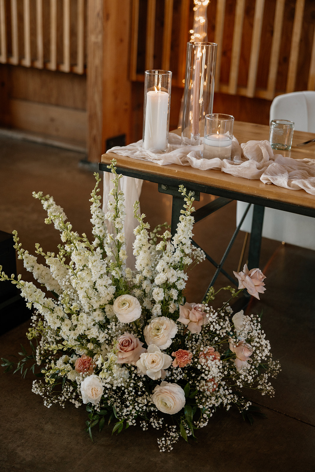 Close-up of a floral arrangement with cream and blush blooms next to soft gauze, candles, and glass details.