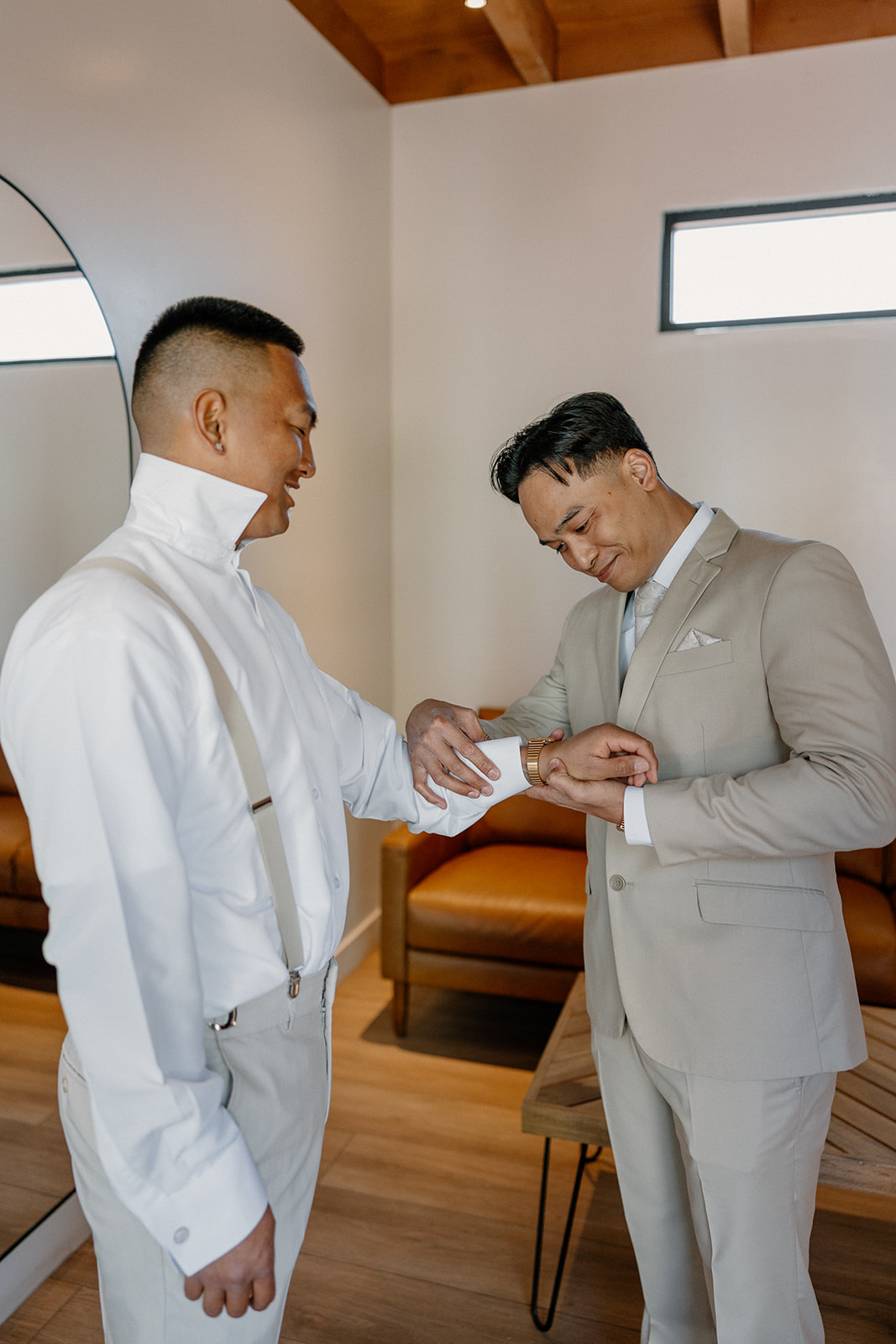 Groom and groomsman sharing a moment while buttoning cuffs in a modern getting-ready suite.