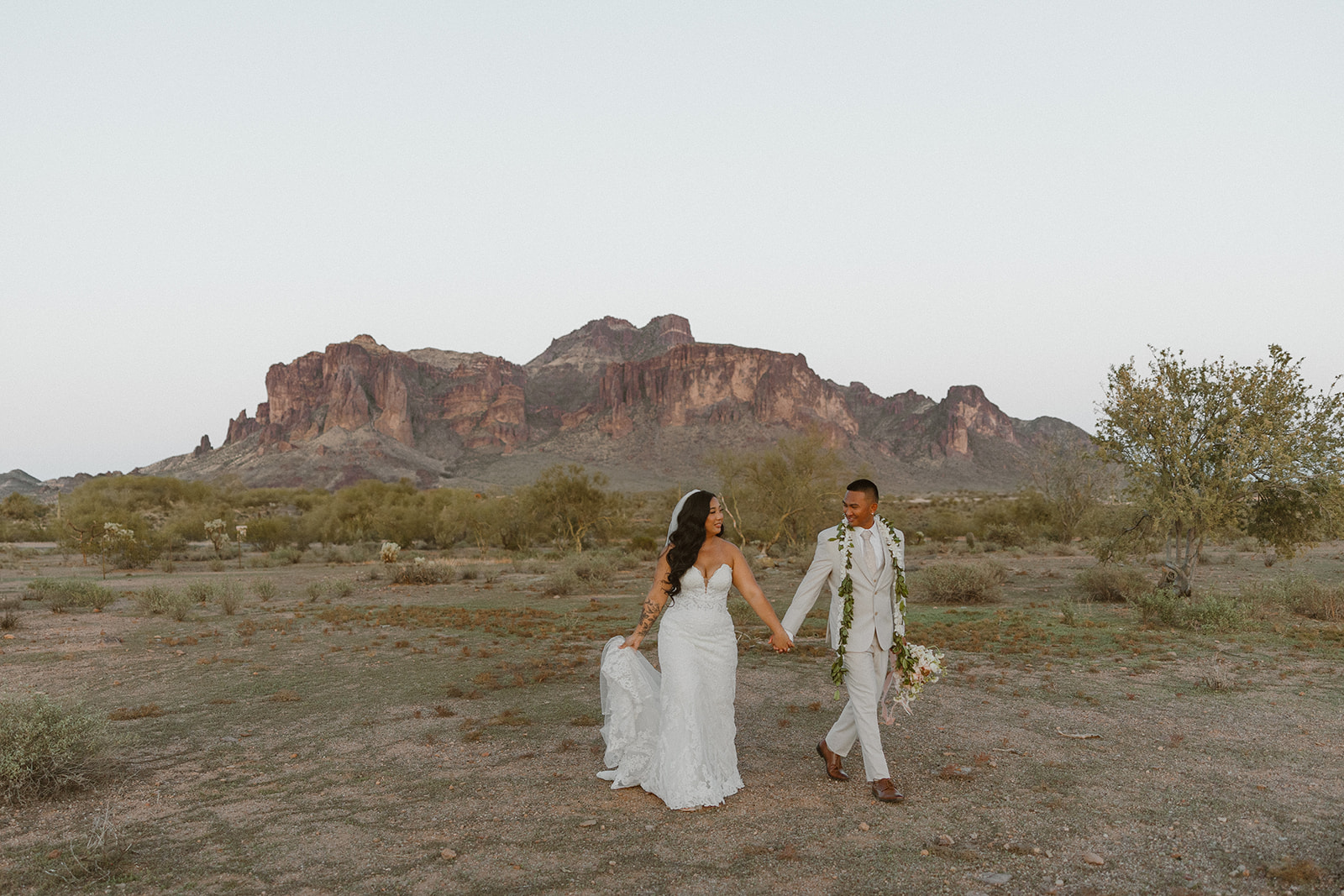 The Paseo wedding couple walking hand-in-hand across the desert at dusk with the Superstition Mountains in the background.
