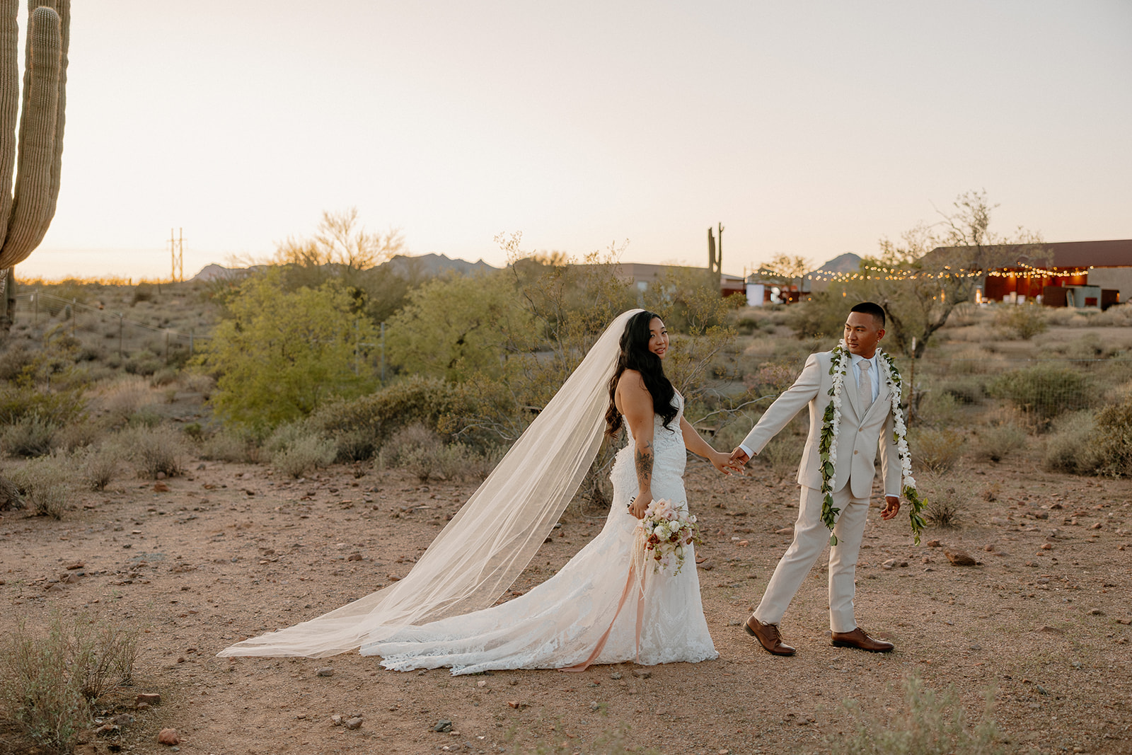 Golden hour portraits of the couple holding hands in the desert, with The Paseo wedding venue lights glowing in the background.