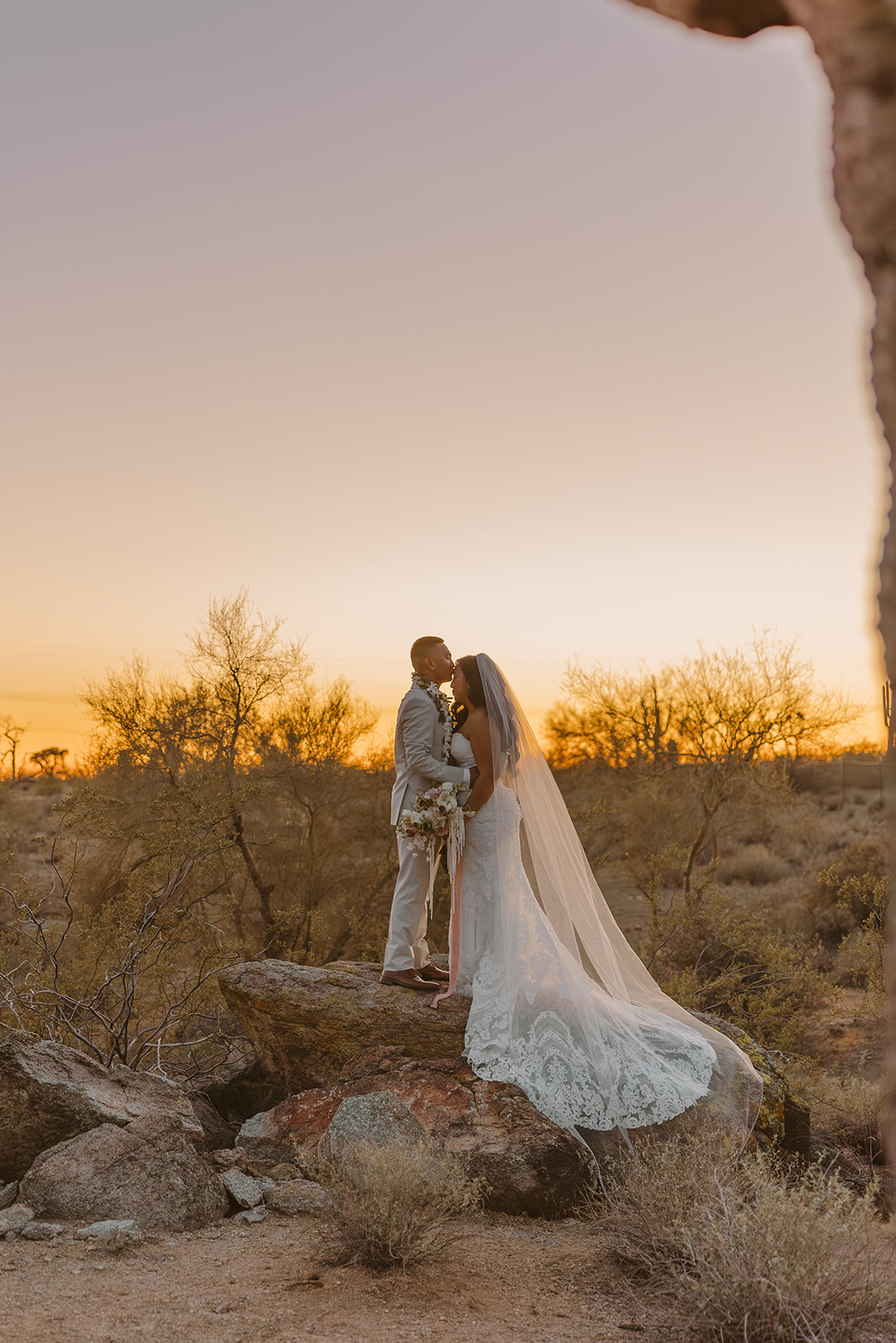 Couple standing on a rock at sunset in full wedding attire, veil flowing, framed by the Arizona desert landscape.