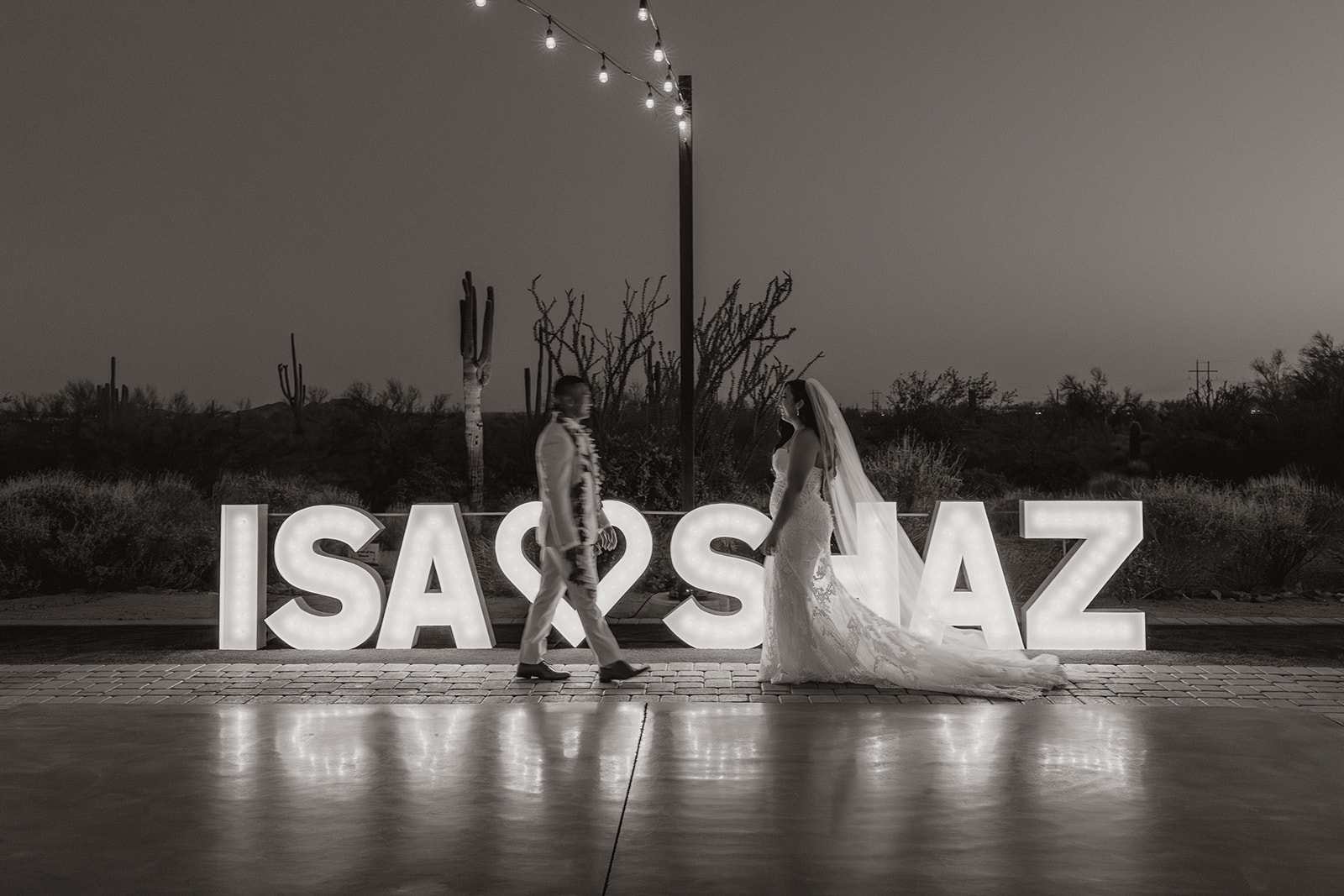 Black and white photo of the couple walking in front of giant light-up marquee letters, desert silhouettes behind them.