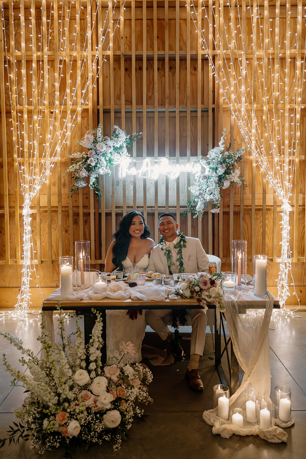 Bride and groom sitting at their candlelit sweetheart table surrounded by flowers and fairy lights, full just-married energy.