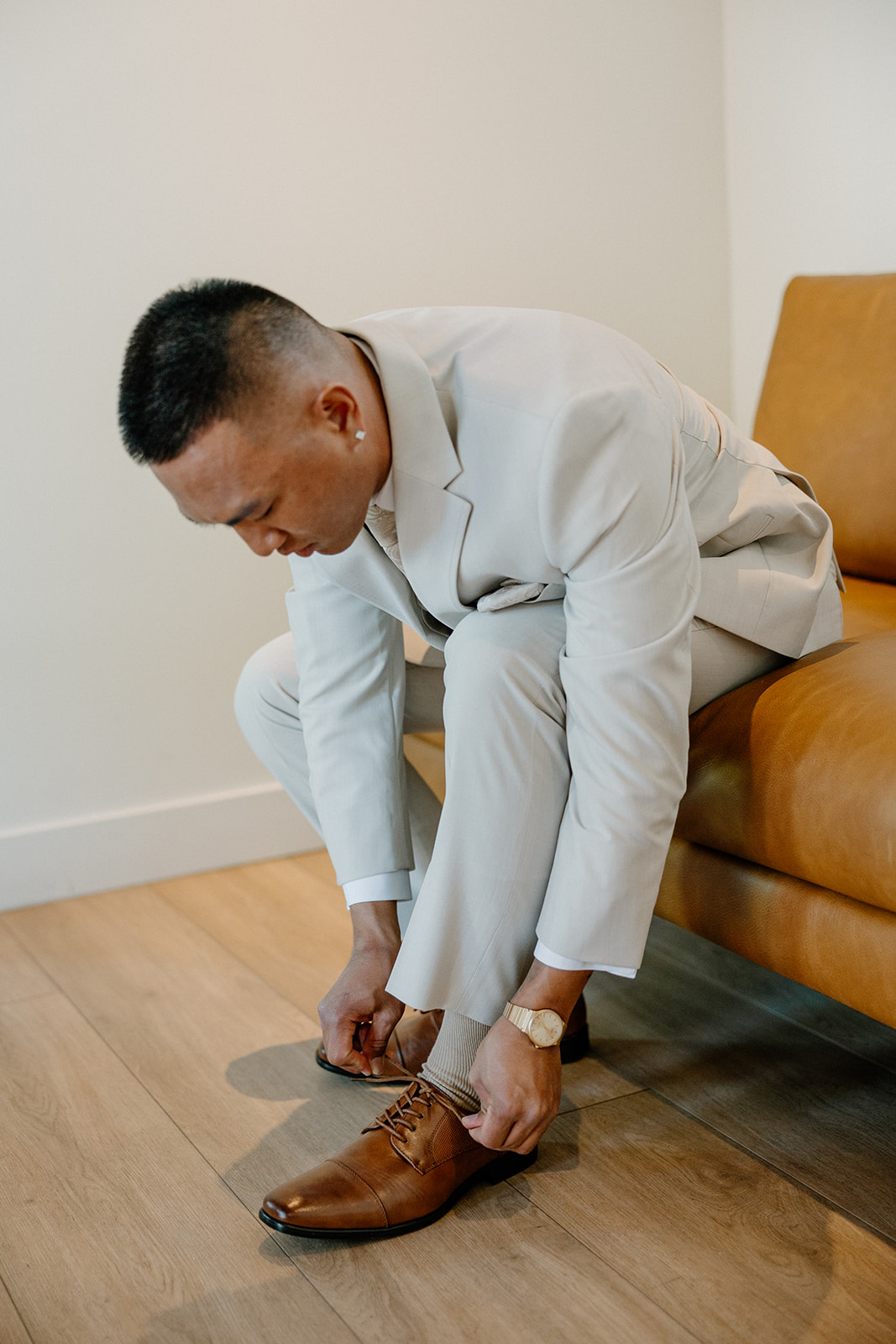 The groom lacing up brown dress shoes on a leather bench in The Paseo wedding prep suite.