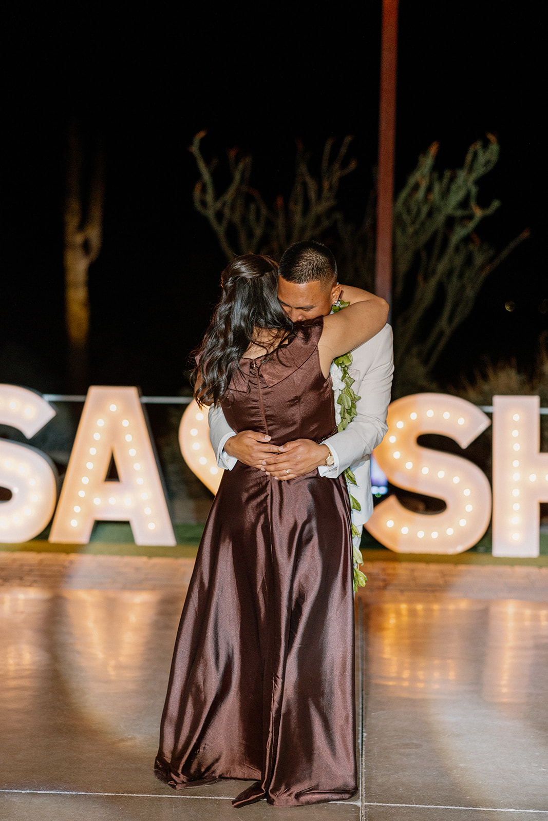 Groom and his mother sharing a warm embrace during their dance, surrounded by soft lighting and marquee glow.