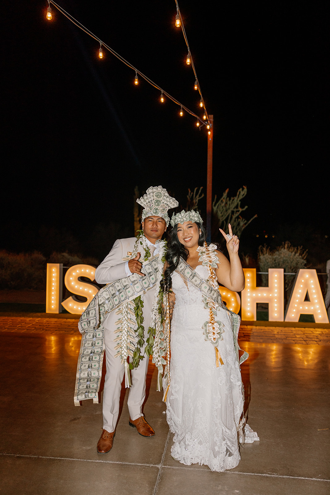Bride and groom rocking money crowns and leis at The Paseo wedding reception under string lights and marquee letters.