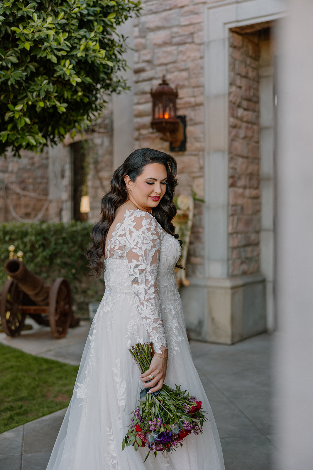 Bride looking over her shoulder while holding a colorful bouquet in front of stone architecture.