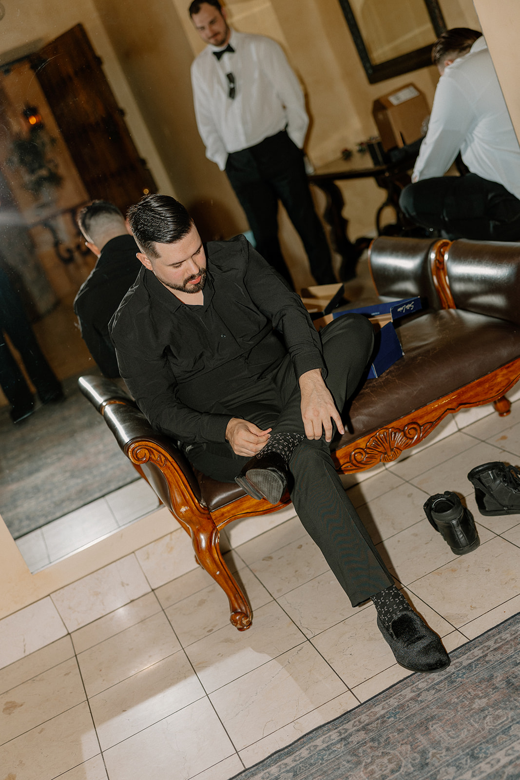 Groom sitting on leather bench adjusting his dress socks while getting ready with his groomsmen.