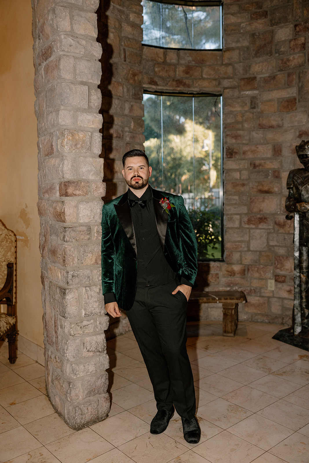 Groom portrait in emerald velvet jacket standing against textured stone walls indoors.