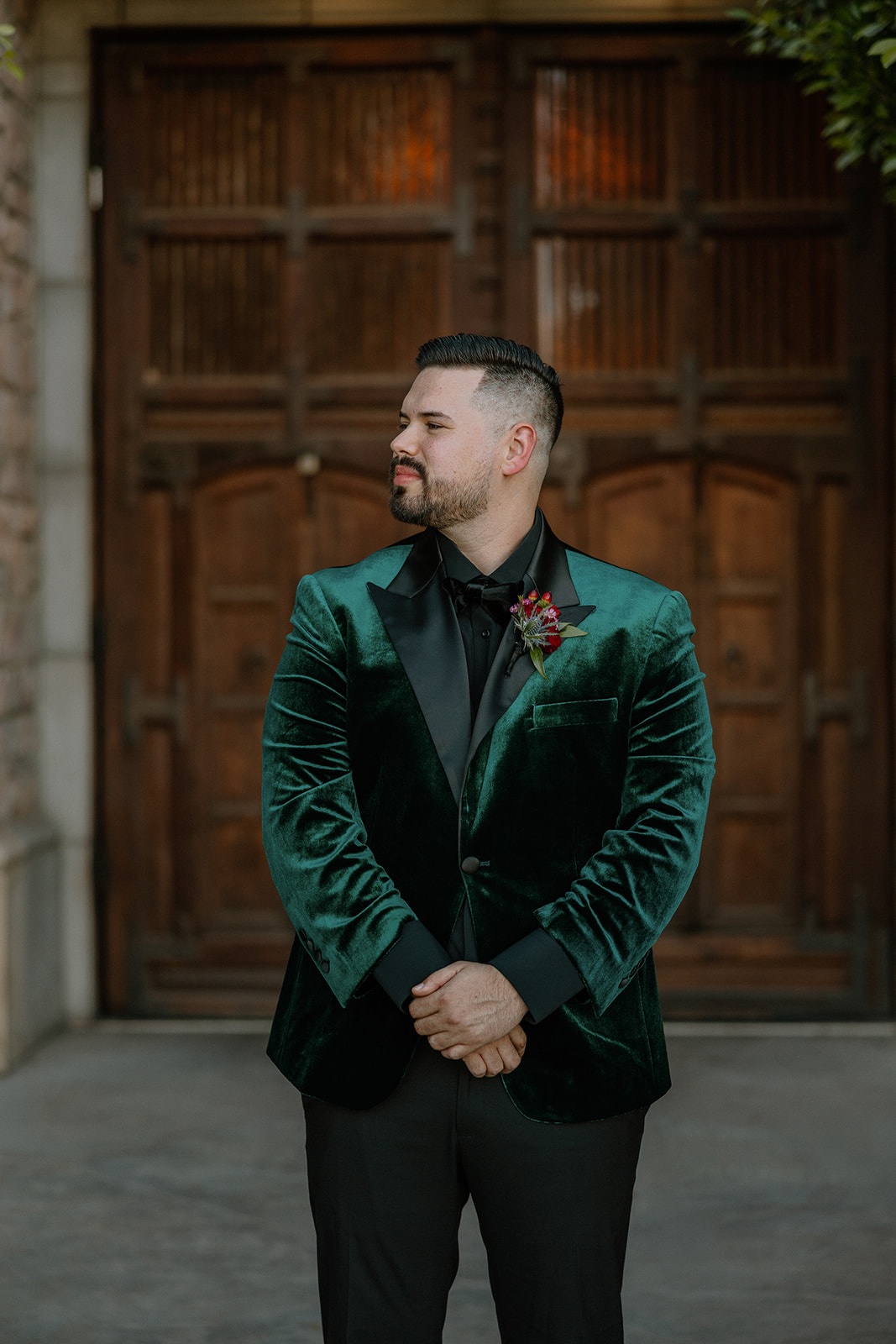 Portrait of groom standing in front of wooden doors in a green velvet tuxedo jacket.