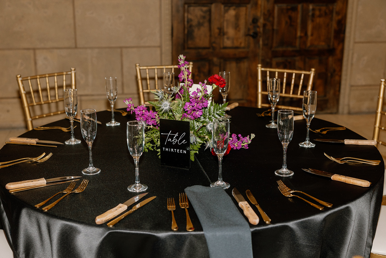 Black reception table with gold flatware, champagne glasses, and bold floral centerpiece against warm stone walls.