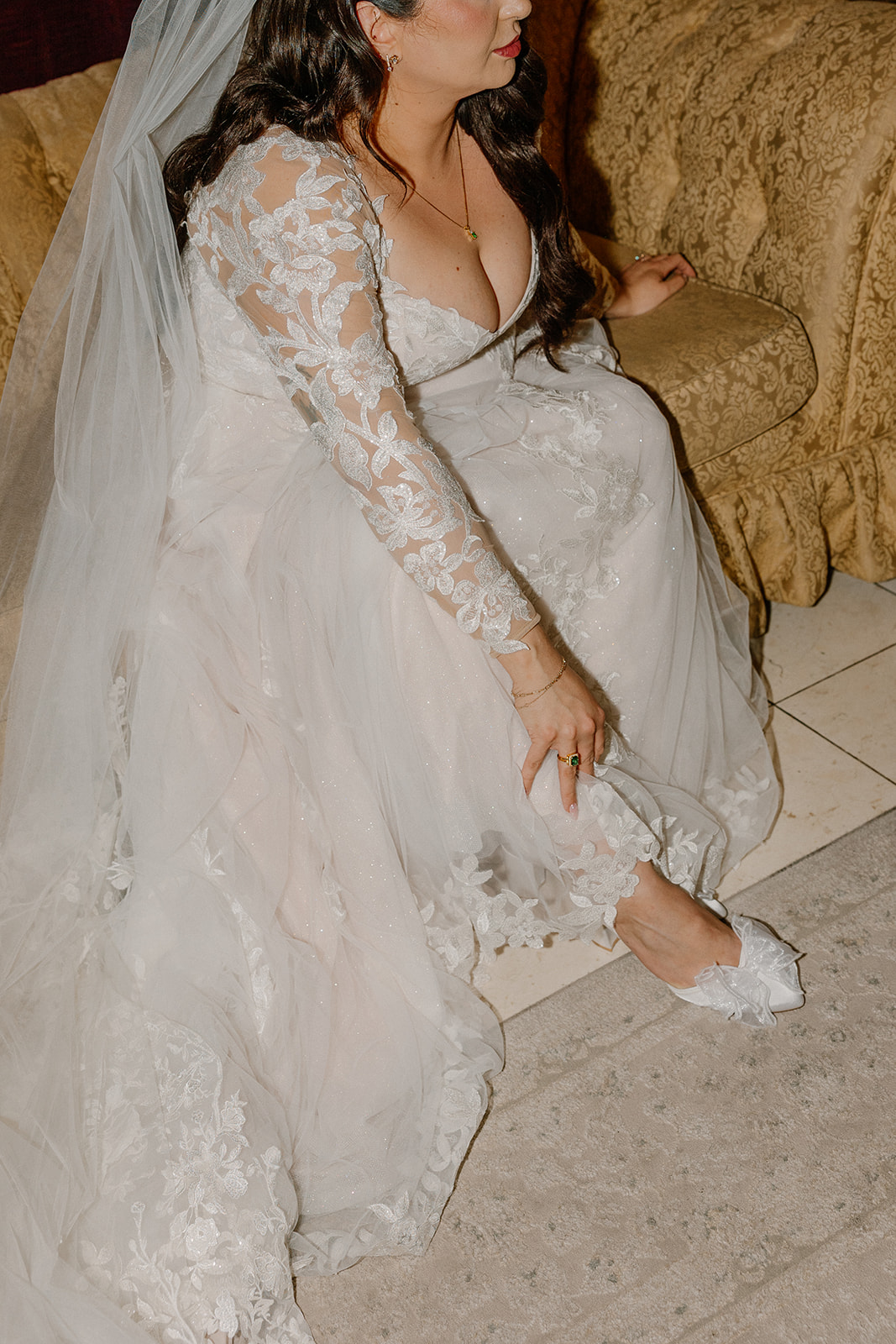 Bride adjusting her wedding heel while seated, showing lace sleeve details and emerald ring.