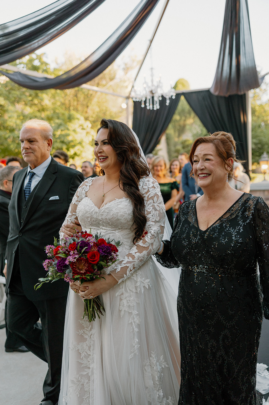 Bride walking down the aisle with her parents, smiling as guests stand.