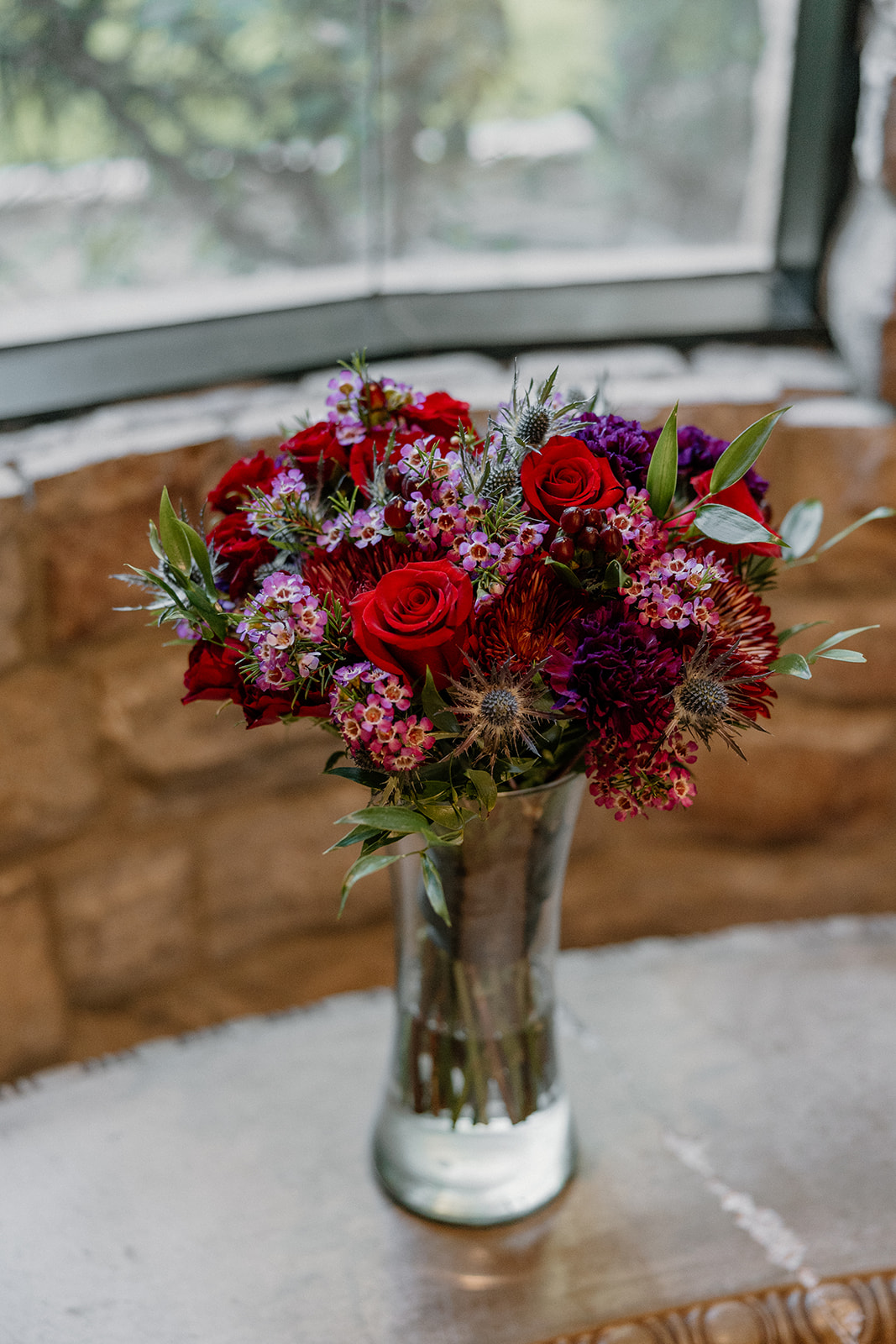 Bold red and berry bridal bouquet in a glass vase with textured greenery and thistle accents.