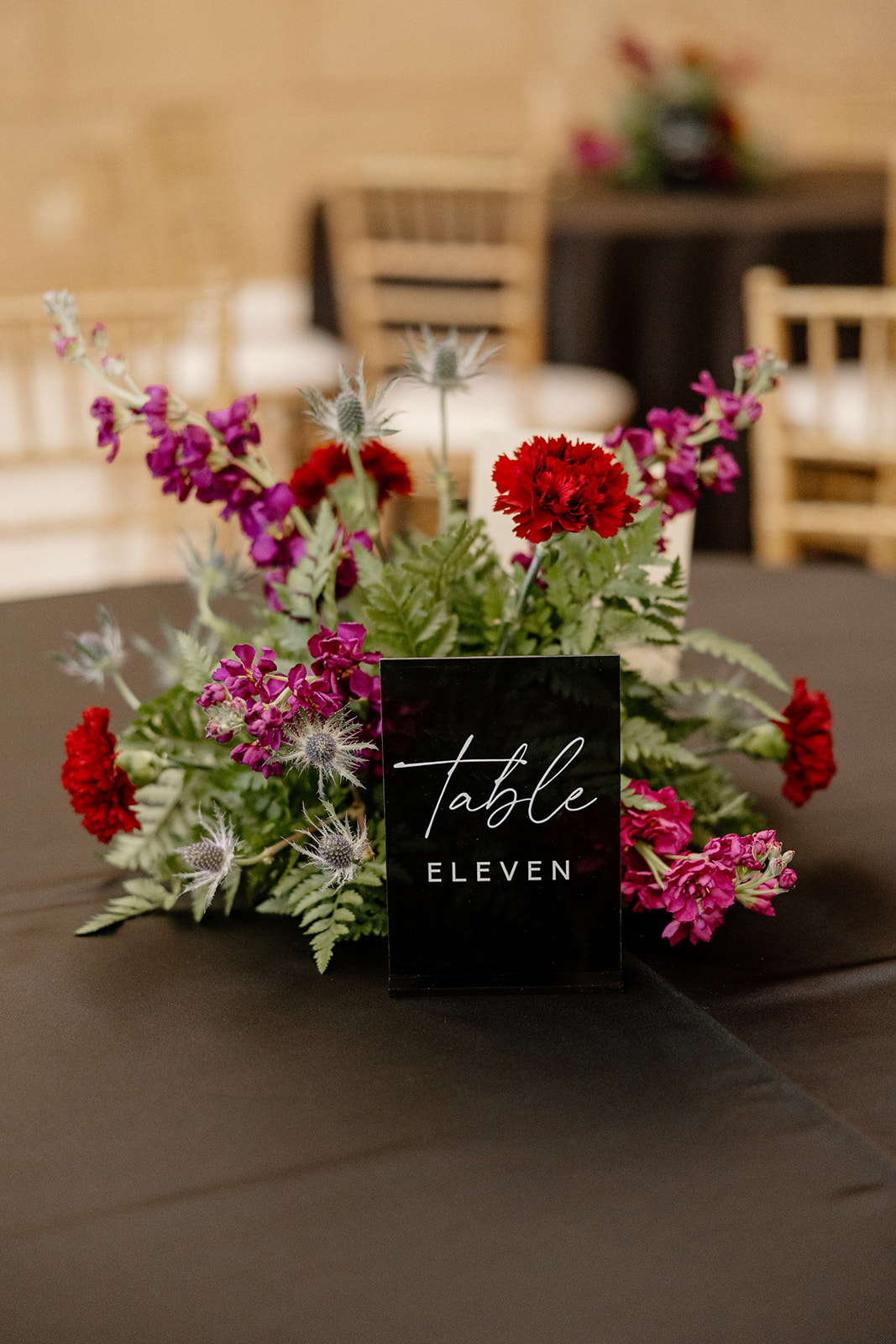 Moody reception centerpiece with red and magenta blooms and modern black table number sign.