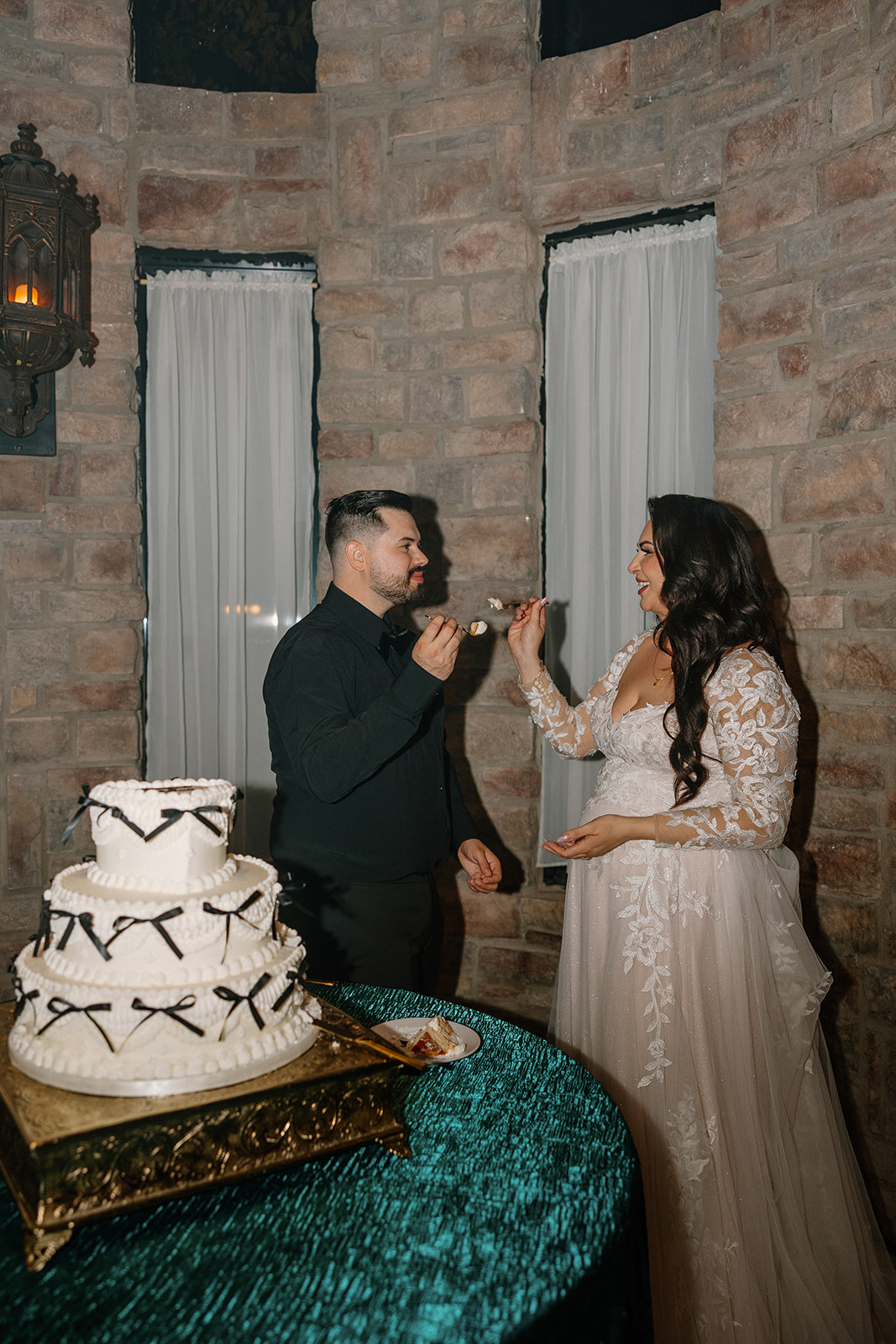 Bride and groom feeding each other cake beside a three-tier wedding cake with black ribbon details.