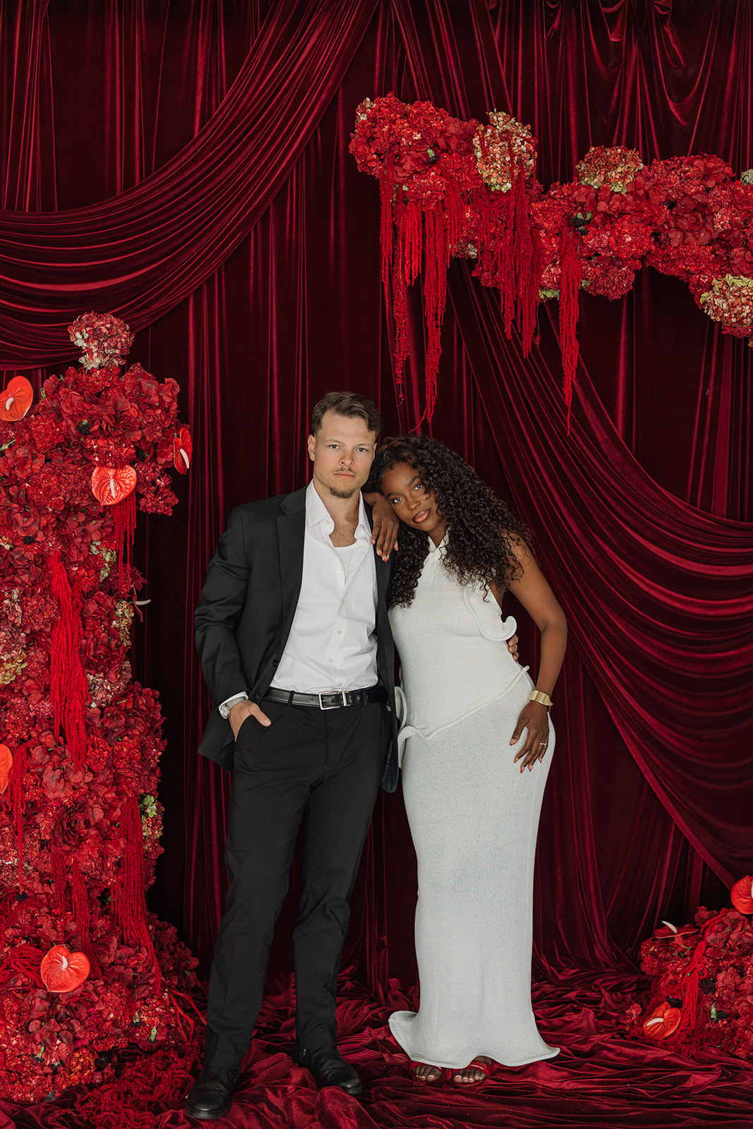 Full-length portrait of the couple standing beneath dramatic red velvet draping and cascading florals, styled in modern formalwear for bold editorial engagement photos.