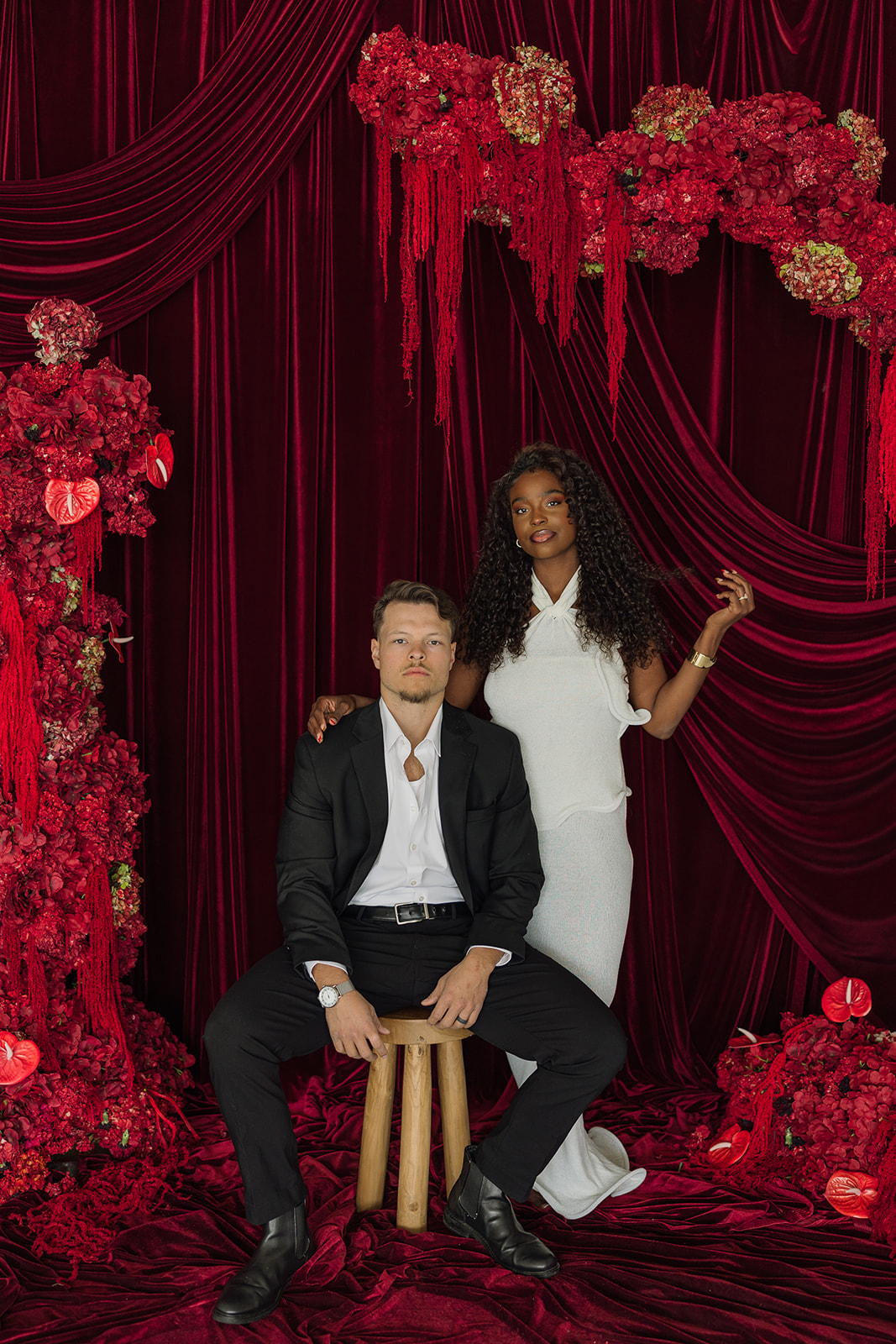 Man sitting on stool as woman stands beside him against dramatic backdrop during editorial engagement photos.