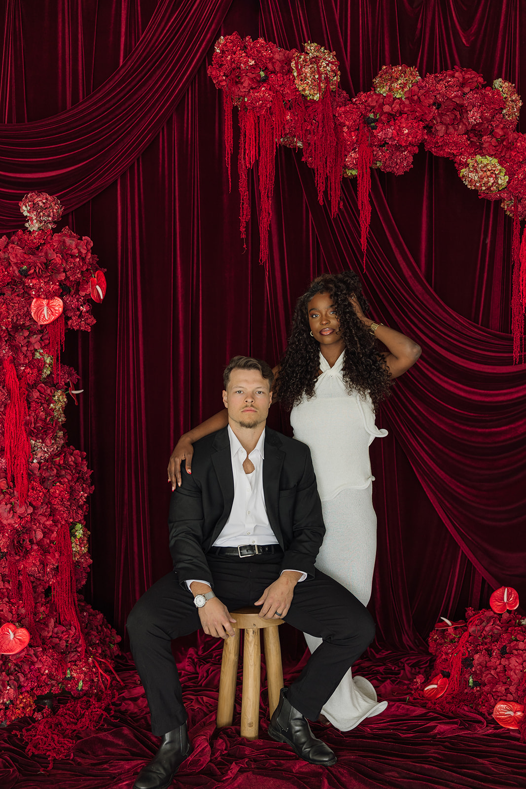 Man sitting on stool as woman stands beside him with her hand in her hair during editorial engagement photos. 