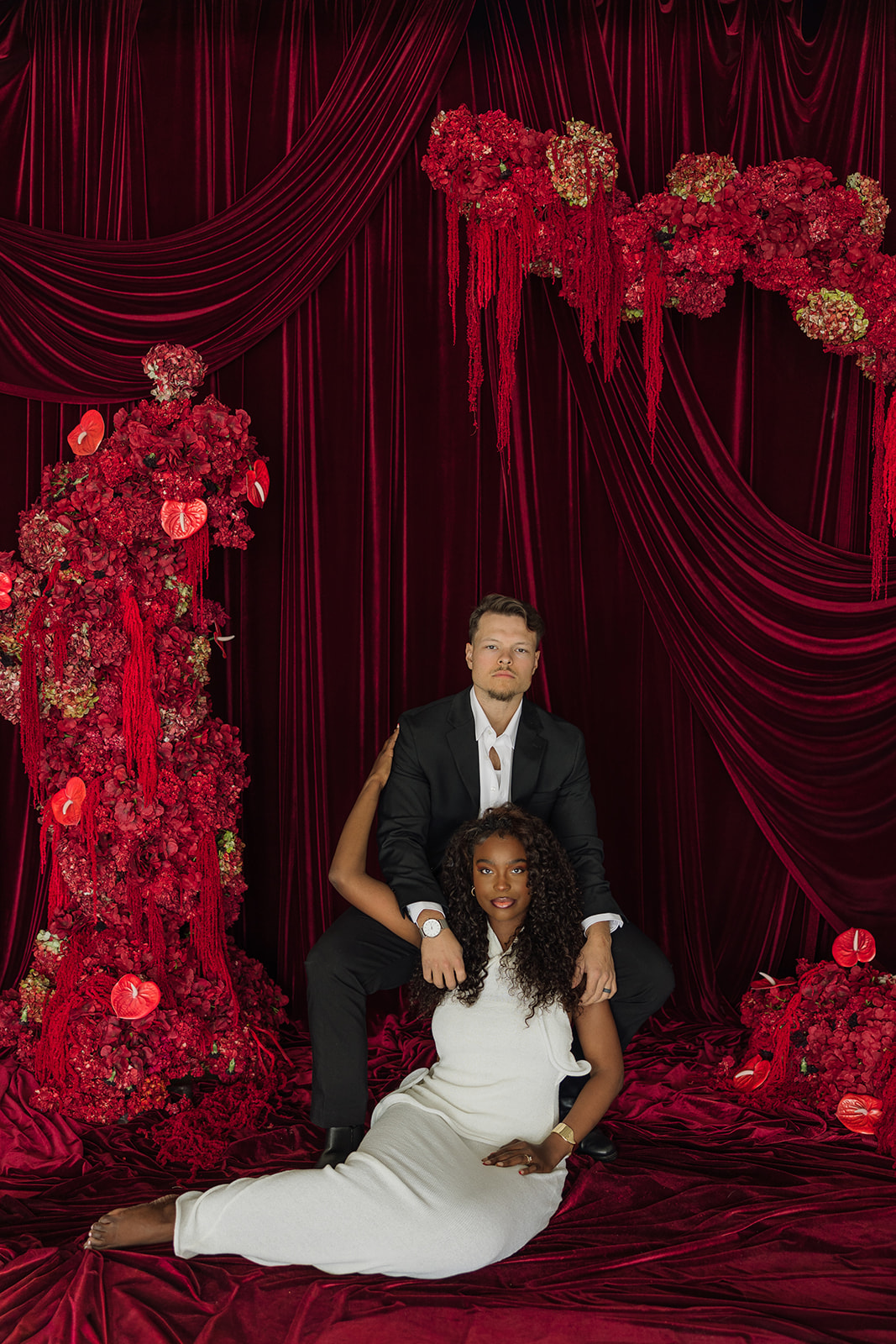 Couple posed against dramatic red velvet drapery and cascading red florals, she seated on the floor in a fitted white dress while he sits behind her in a black suit for bold editorial engagement photos.