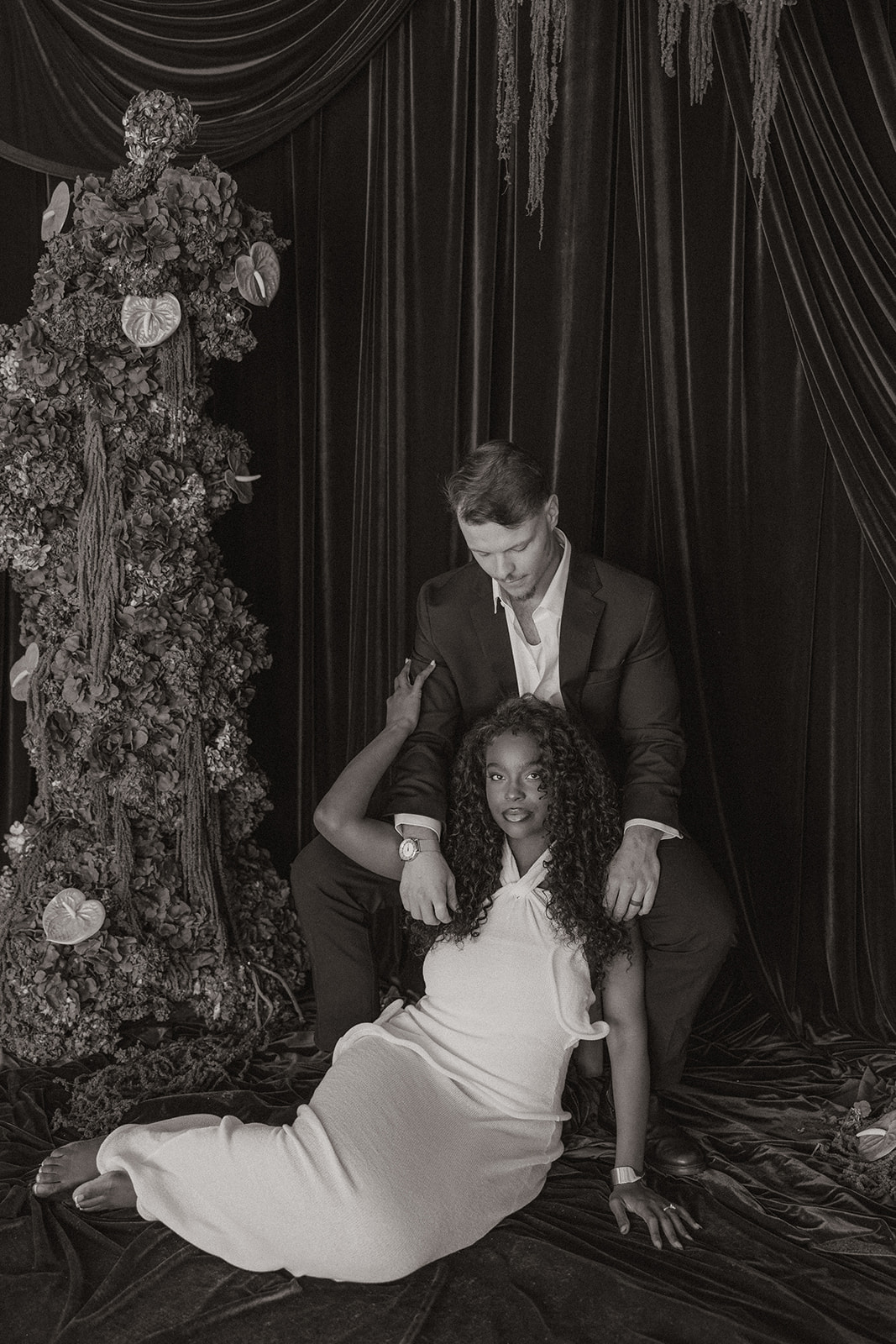 Monochrome full-body portrait of the couple posed against velvet curtains and dramatic floral textures, blending classic styling with editorial engagement photos.
