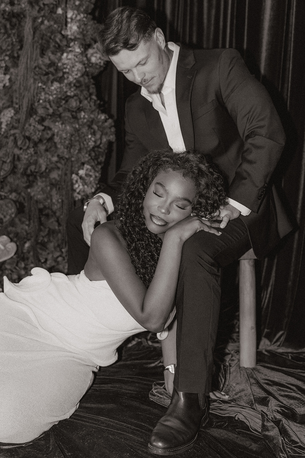 Black and white portrait of the couple in an intimate pose, she resting her head on his knee while he looks down, styled as timeless editorial engagement photos.