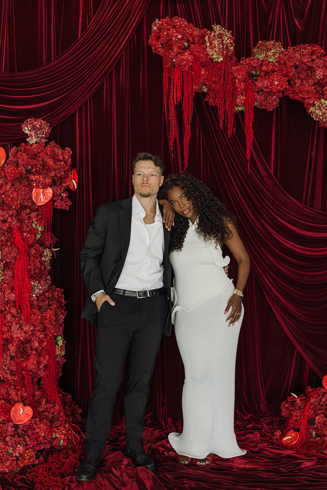 Full-length portrait of the couple standing beneath red floral installations and velvet draping, styled in modern black and white formalwear.