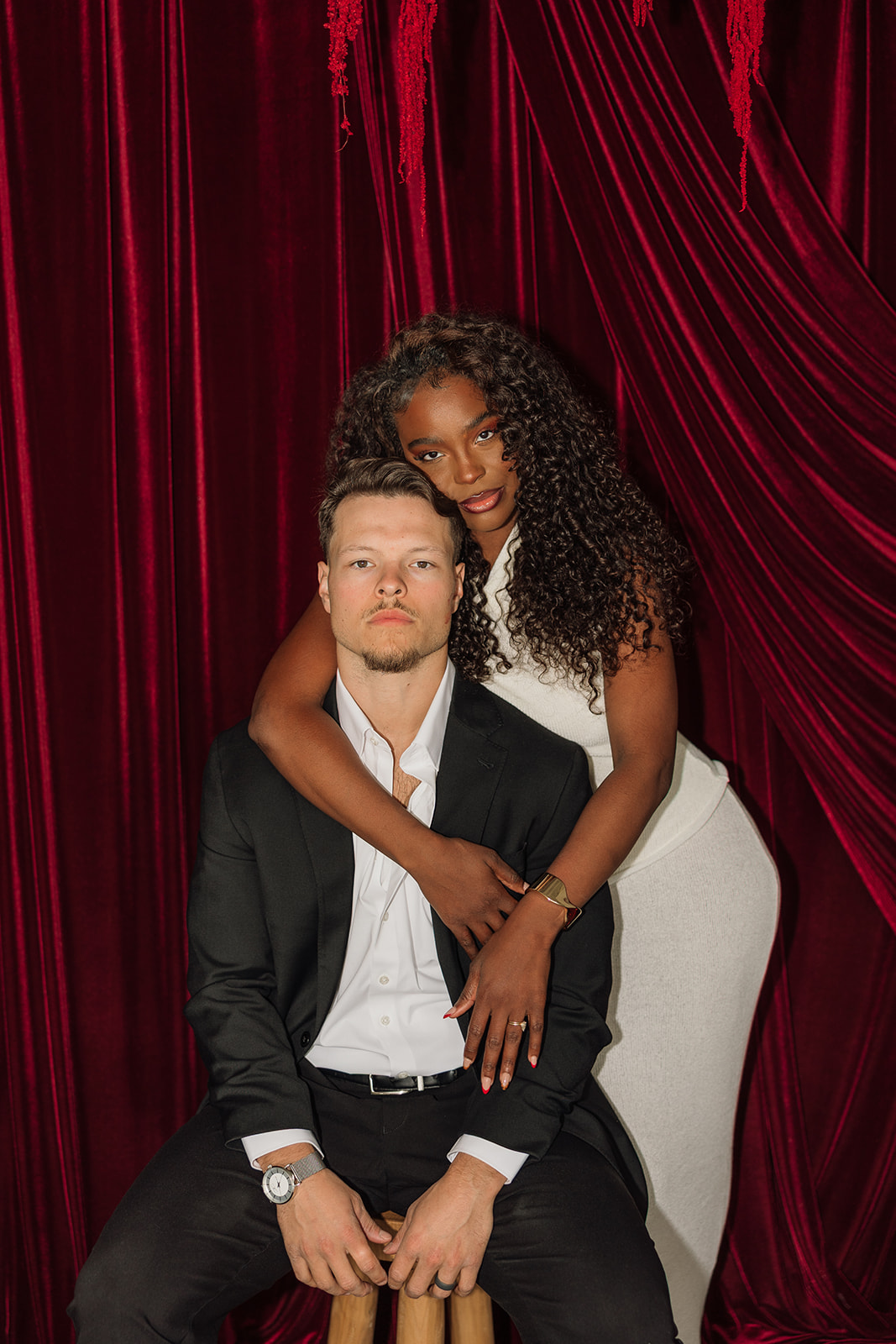 Intimate seated portrait with her arms wrapped around his shoulders as they look toward the camera, framed by deep red velvet for elevated editorial engagement photos.