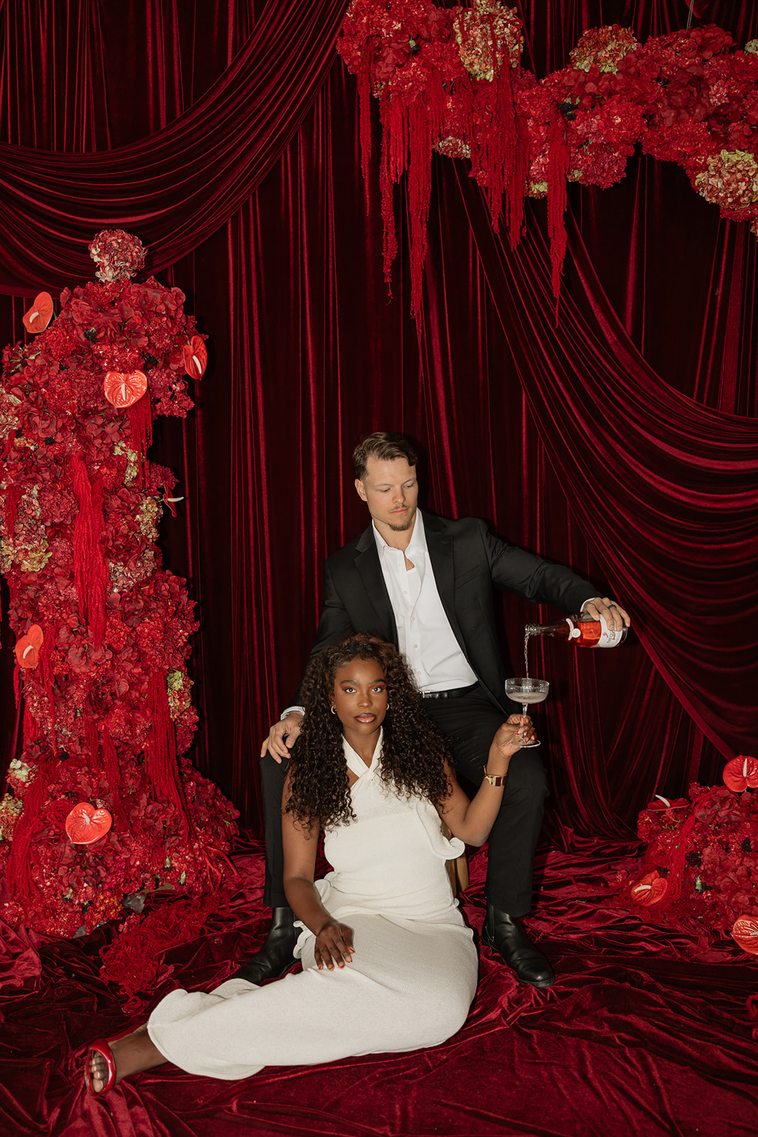 Couple styled in black and white formalwear with champagne and statement red floral installations, creating a high-fashion editorial engagement photos look.