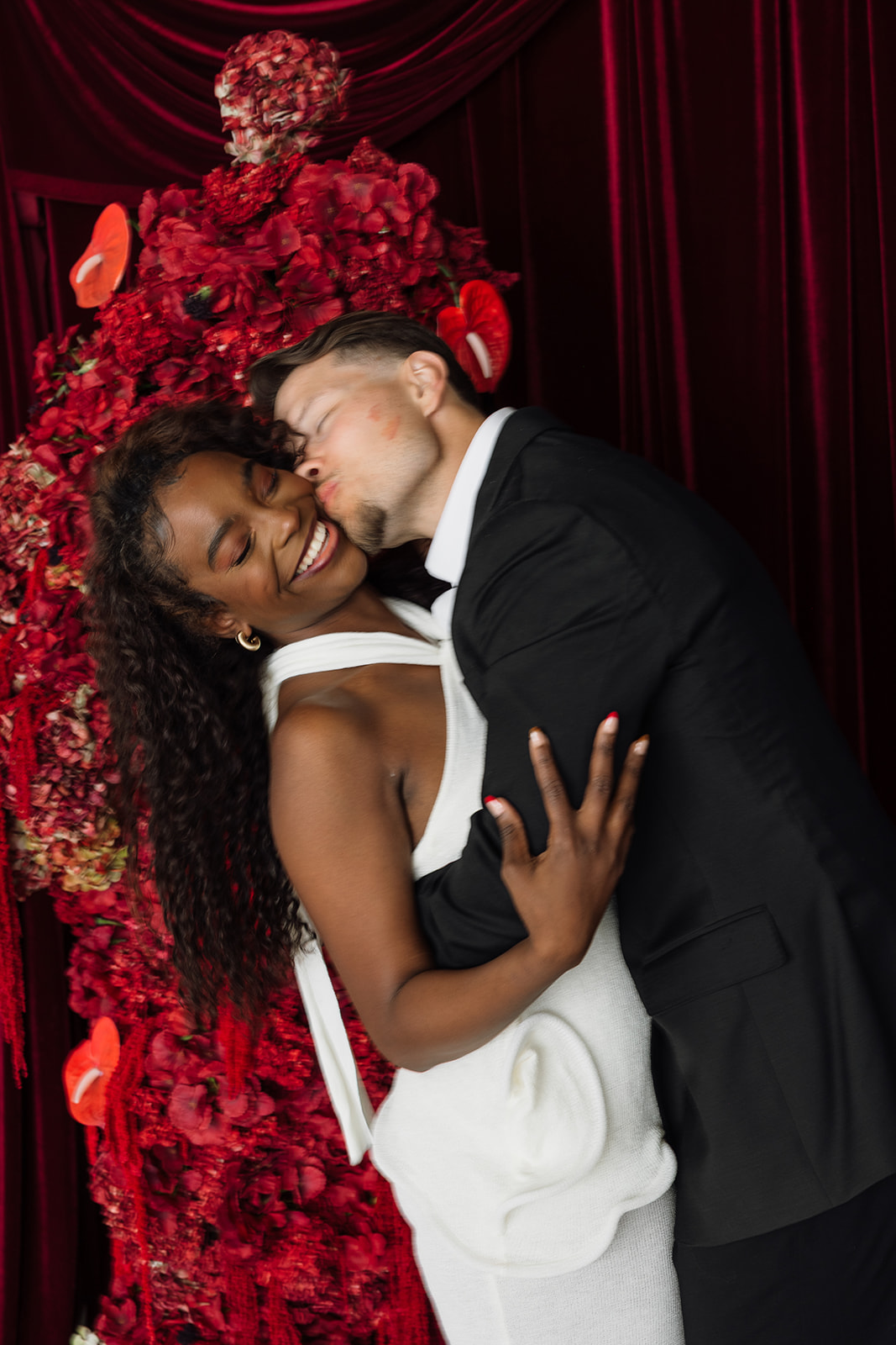 Man kisses woman on cheek while she laughs with remnants of red lipstick on his cheek